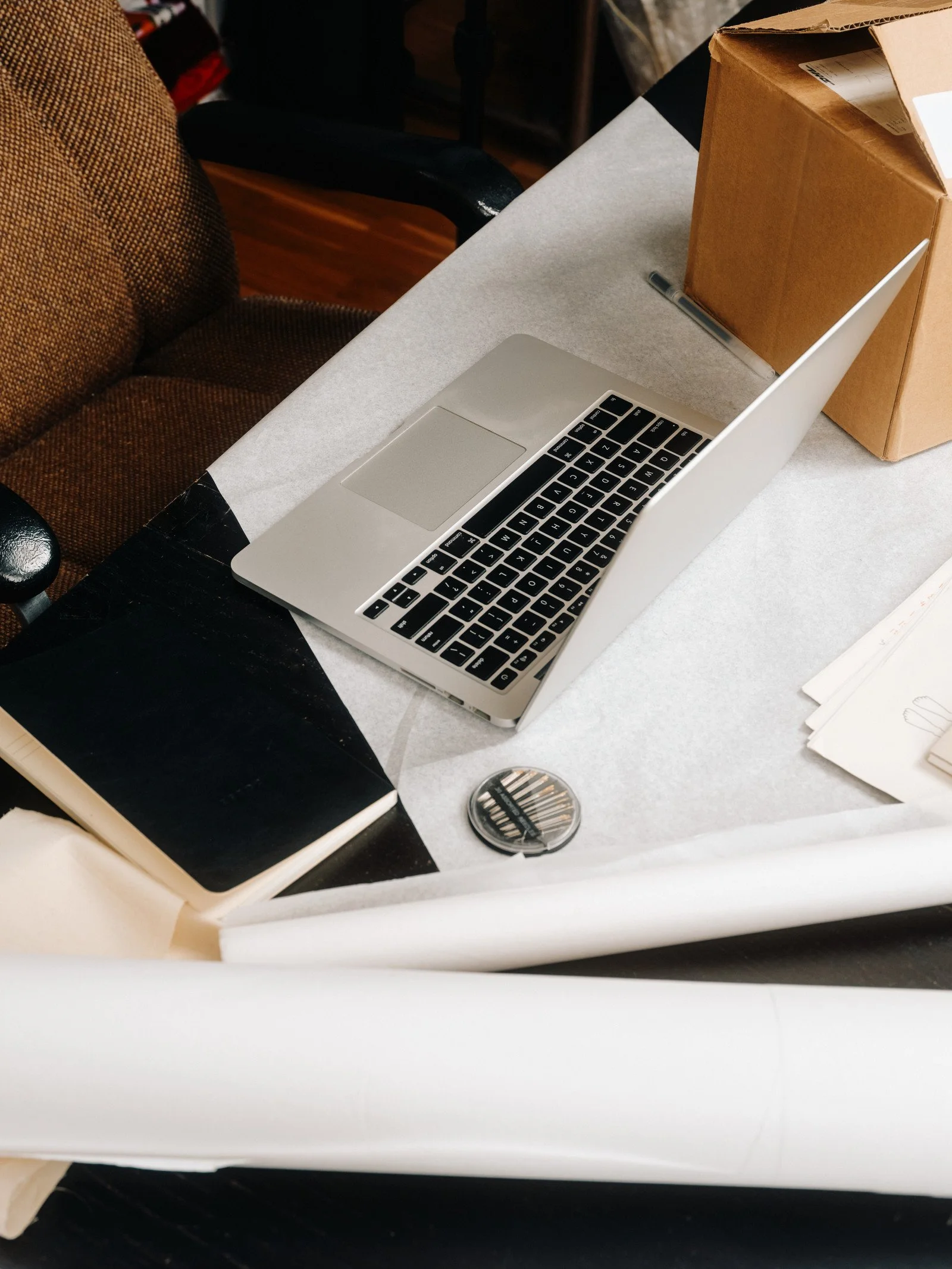 A workspace with a laptop, brown cardboard box, black notebook, pen, and a round container on a white table.