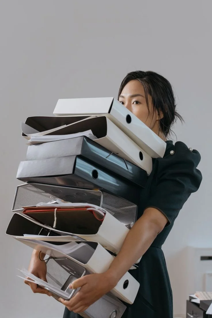 A woman with black hair bundled back, wearing a dark short-sleeved dress, holding a large stack of binders, folders, and papers in an office setting.