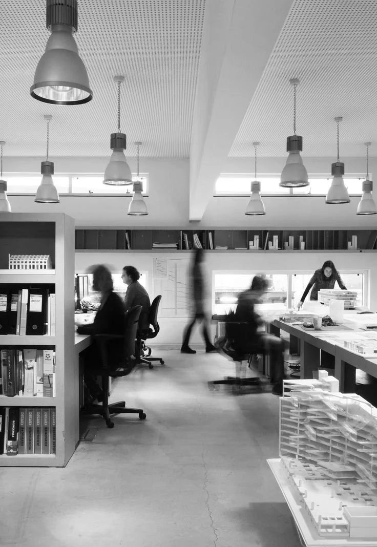 A black and white photo of an office space with several people working at desks. There are hanging ceiling lights, bookshelves, and a large window in the background. Some people are seated at desks, while others are walking or standing.
