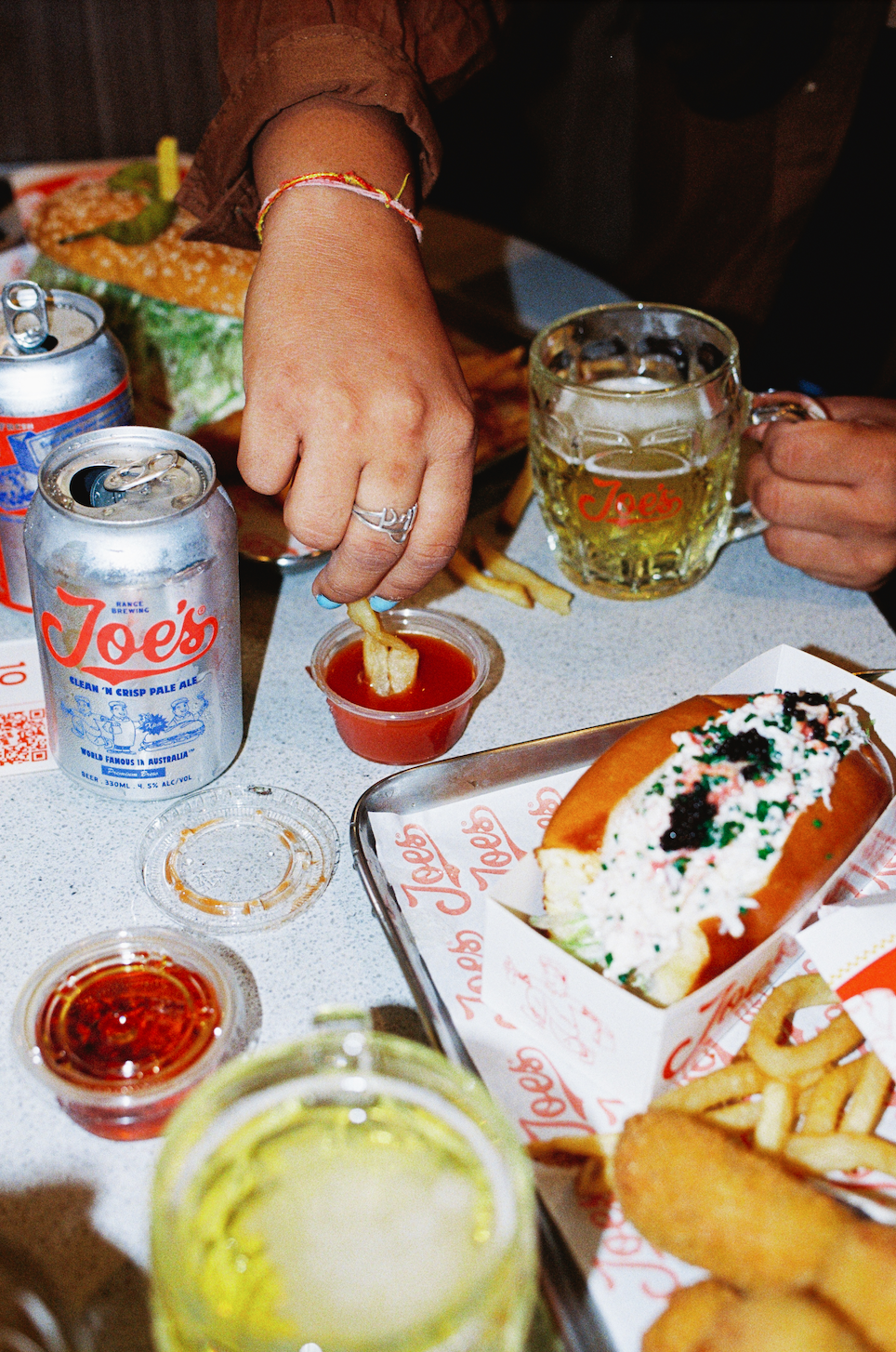 People eating at a table with drinks, hot dog topped with mayonnaise and relish, fries, onion rings, and dipping sauces. One person dips fries into ketchup.
