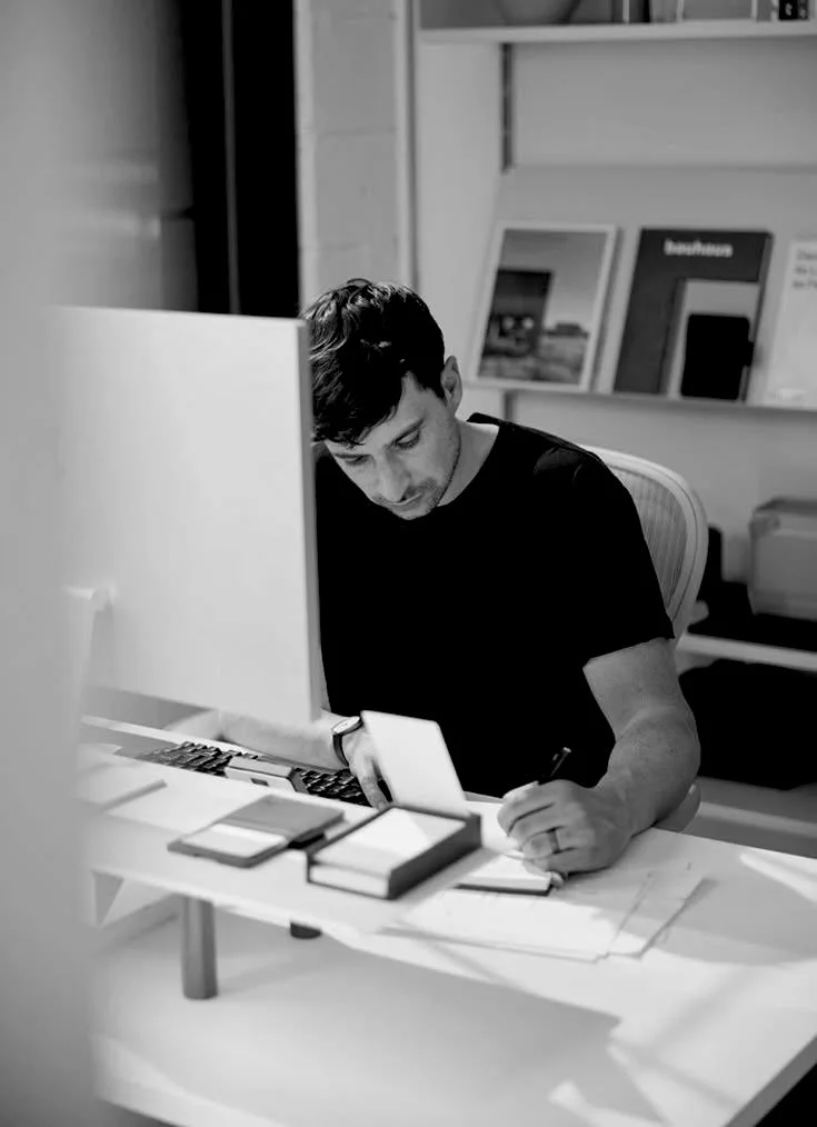 A young man working at a desk, writing on papers, with books and computer equipment around him in an office setting.