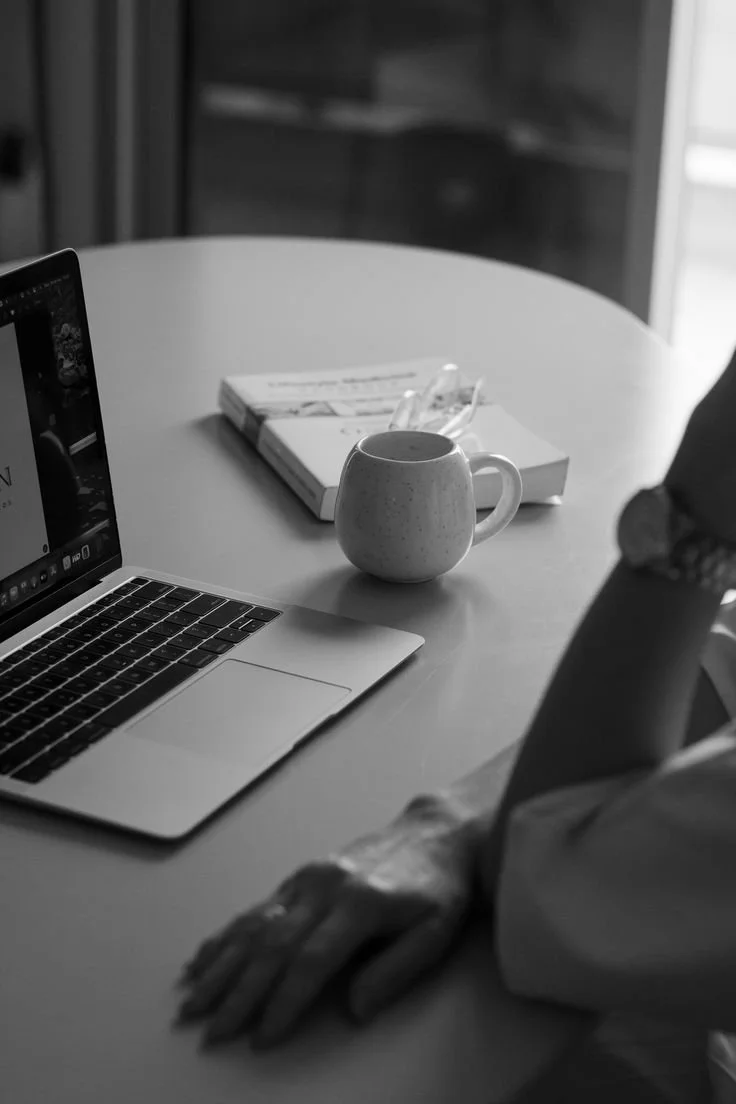 A person sitting at a round table with a laptop, a mug, and a magazine or book with a clear pen on top. The person has their arms resting on the table, and only part of their body is visible.