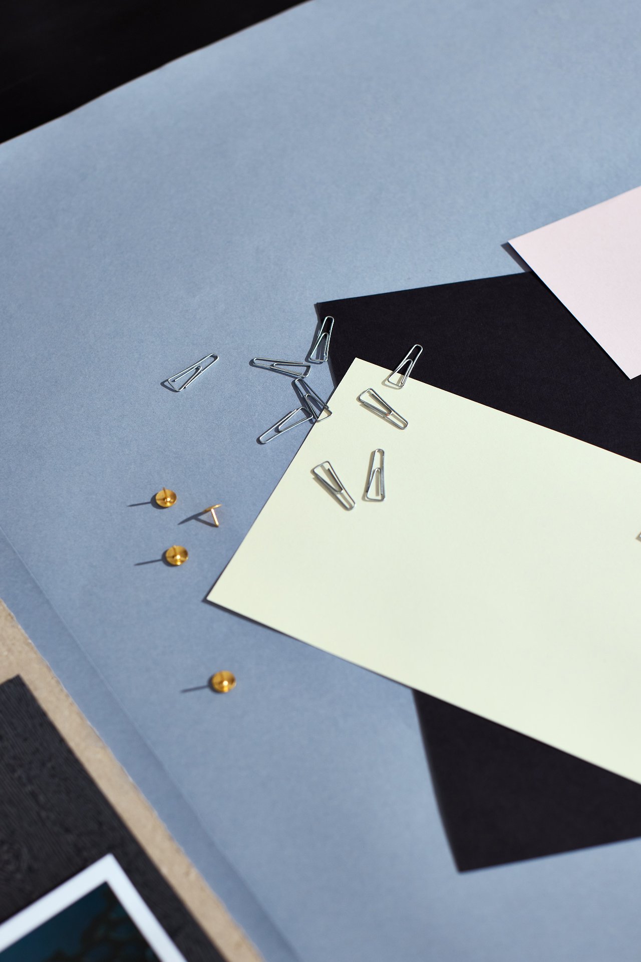A flat lay of decorative paper in light blue, pale yellow, and white, with scattered silver paper clips and small gold push pins on a dark surface.
