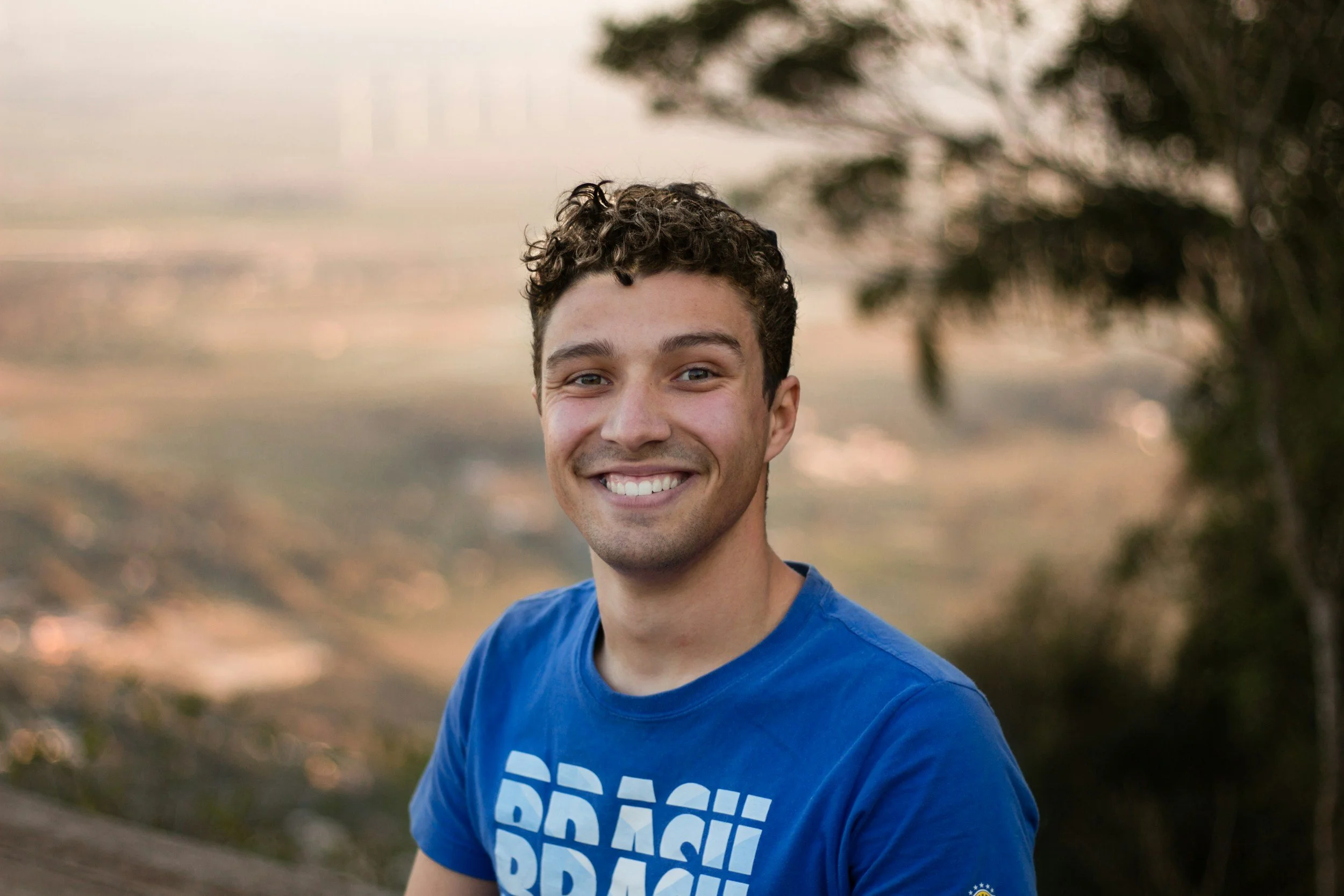 A smiling young man with curly hair wearing a blue T-shirt, standing outdoors with a blurred natural landscape background.