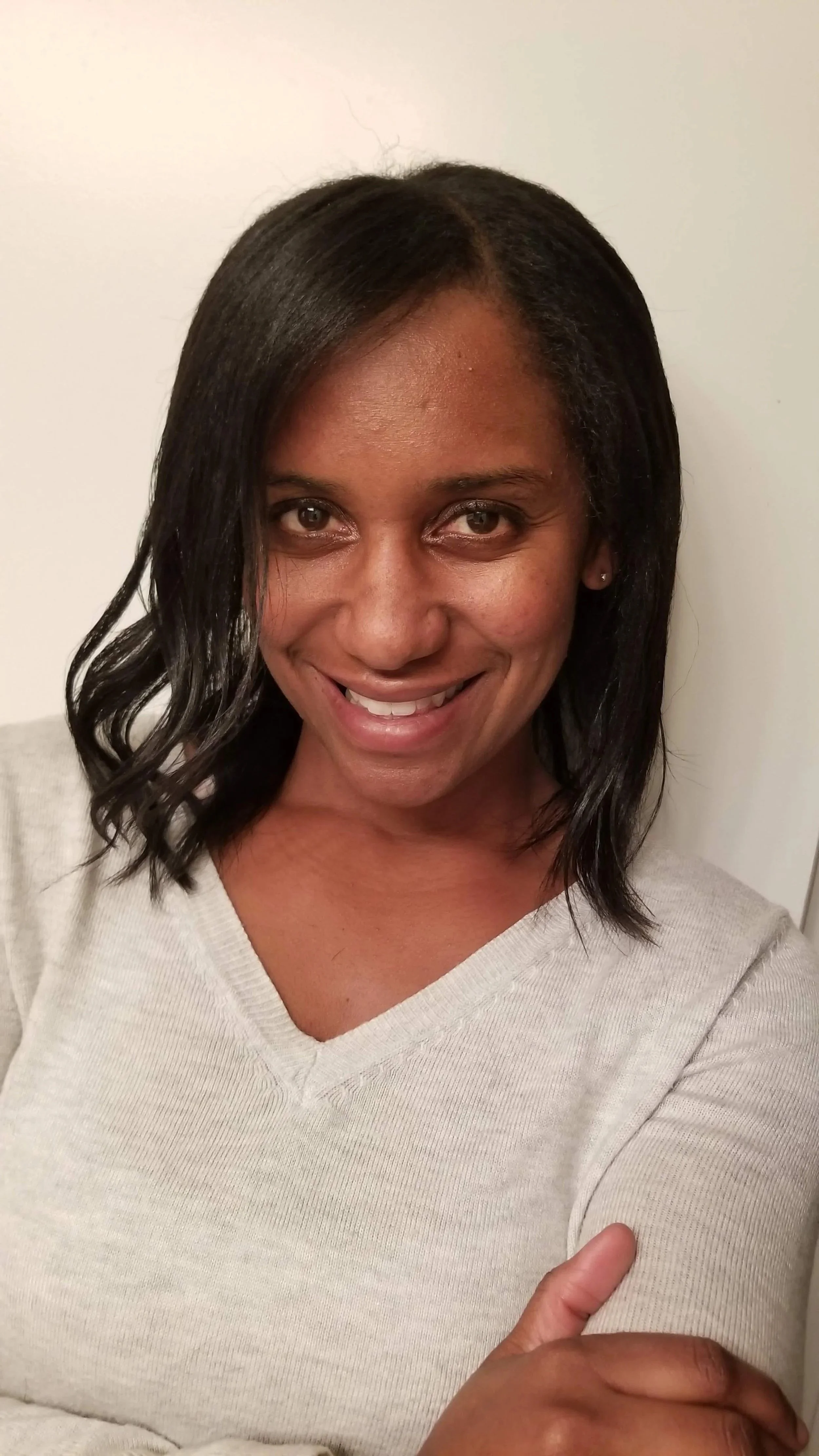 Close-up of a woman with medium-dark skin and black wavy shoulder-length hair, smiling in front of a plain white wall.