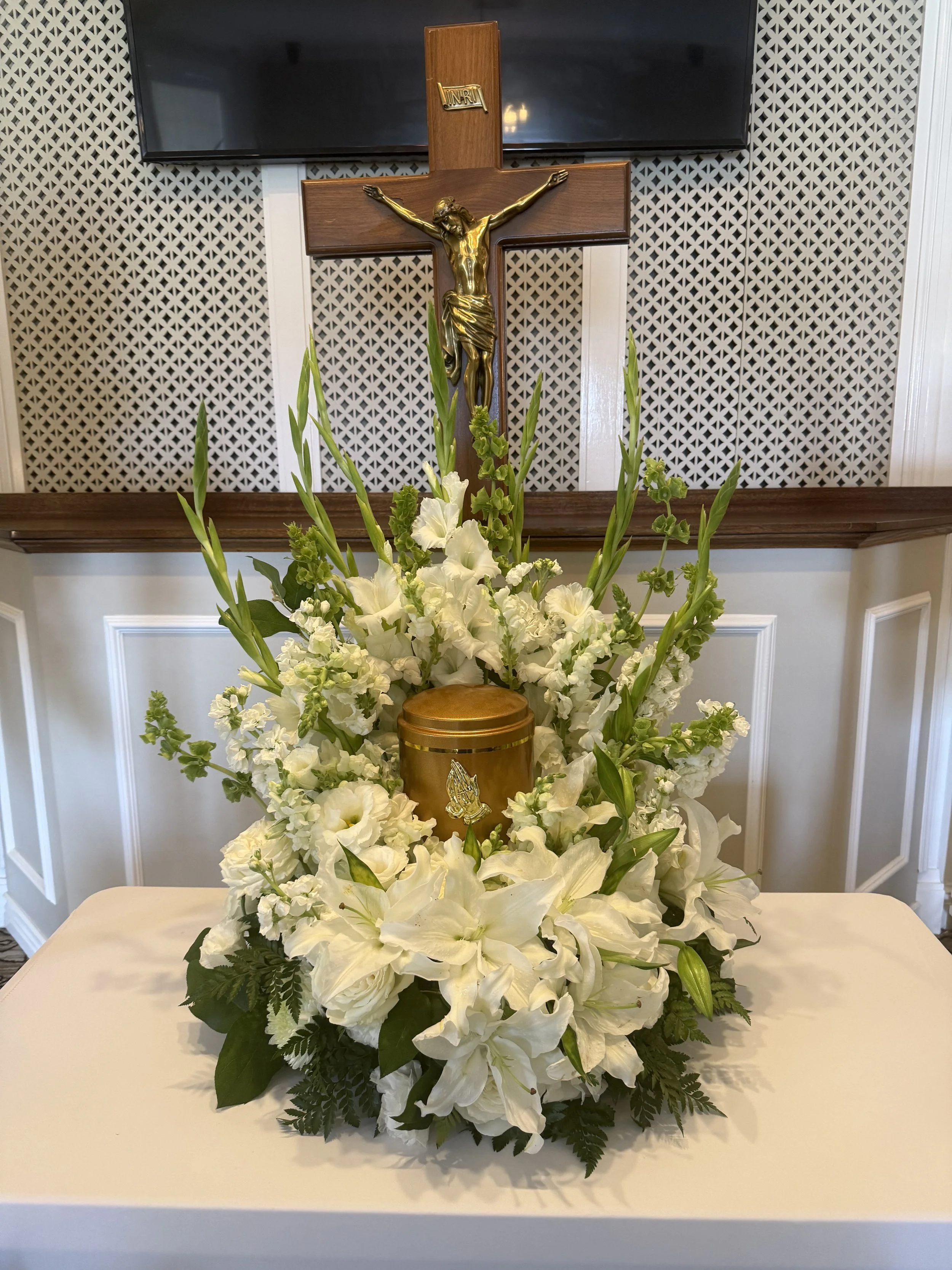 A wooden crucifix with a gold-colored Jesus figure and an INRI sign, placed above a bouquet of white flowers, on a table in front of a patterned wall and a television.