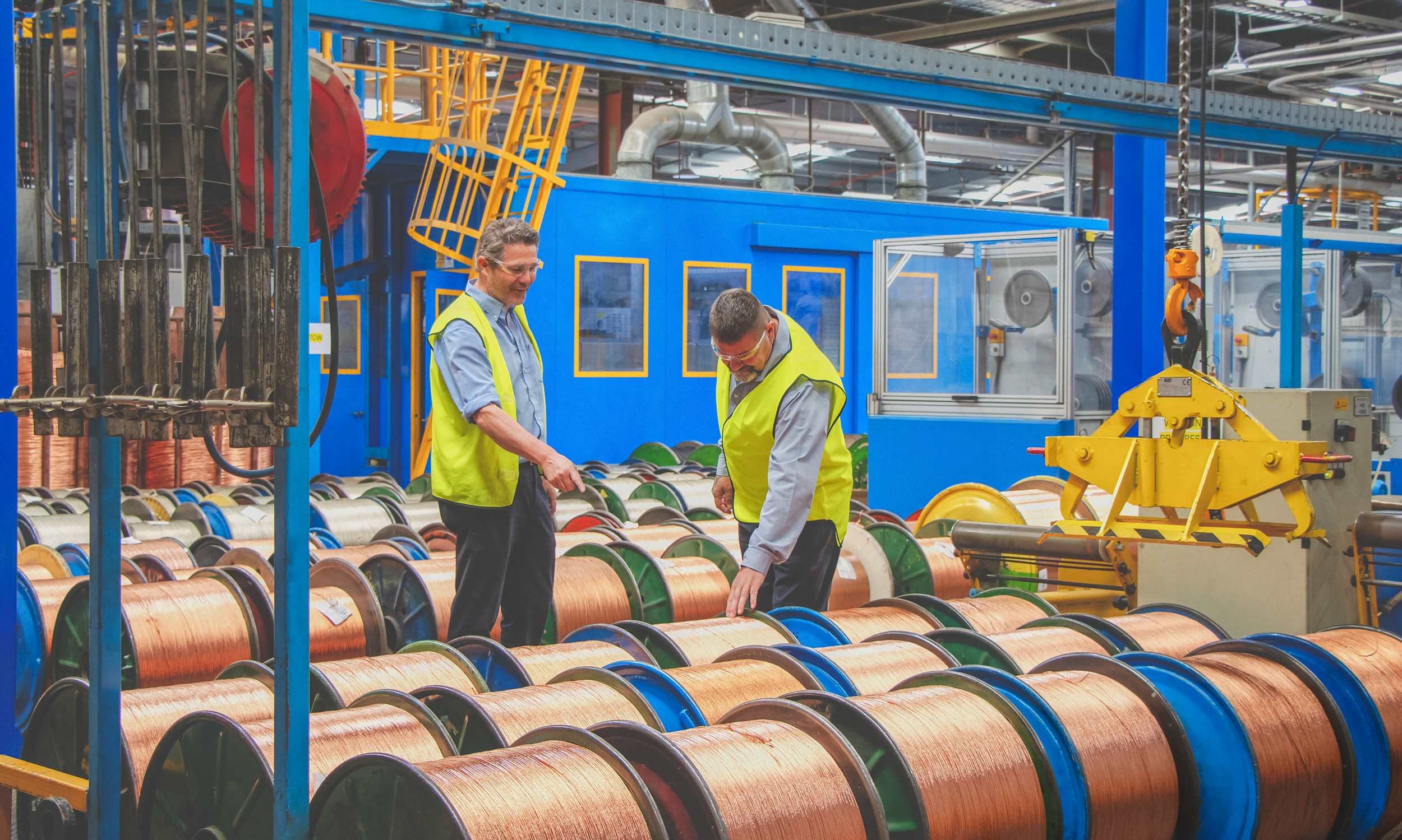 Two workers in safety vests inspecting copper wires on spools in an industrial manufacturing facility.