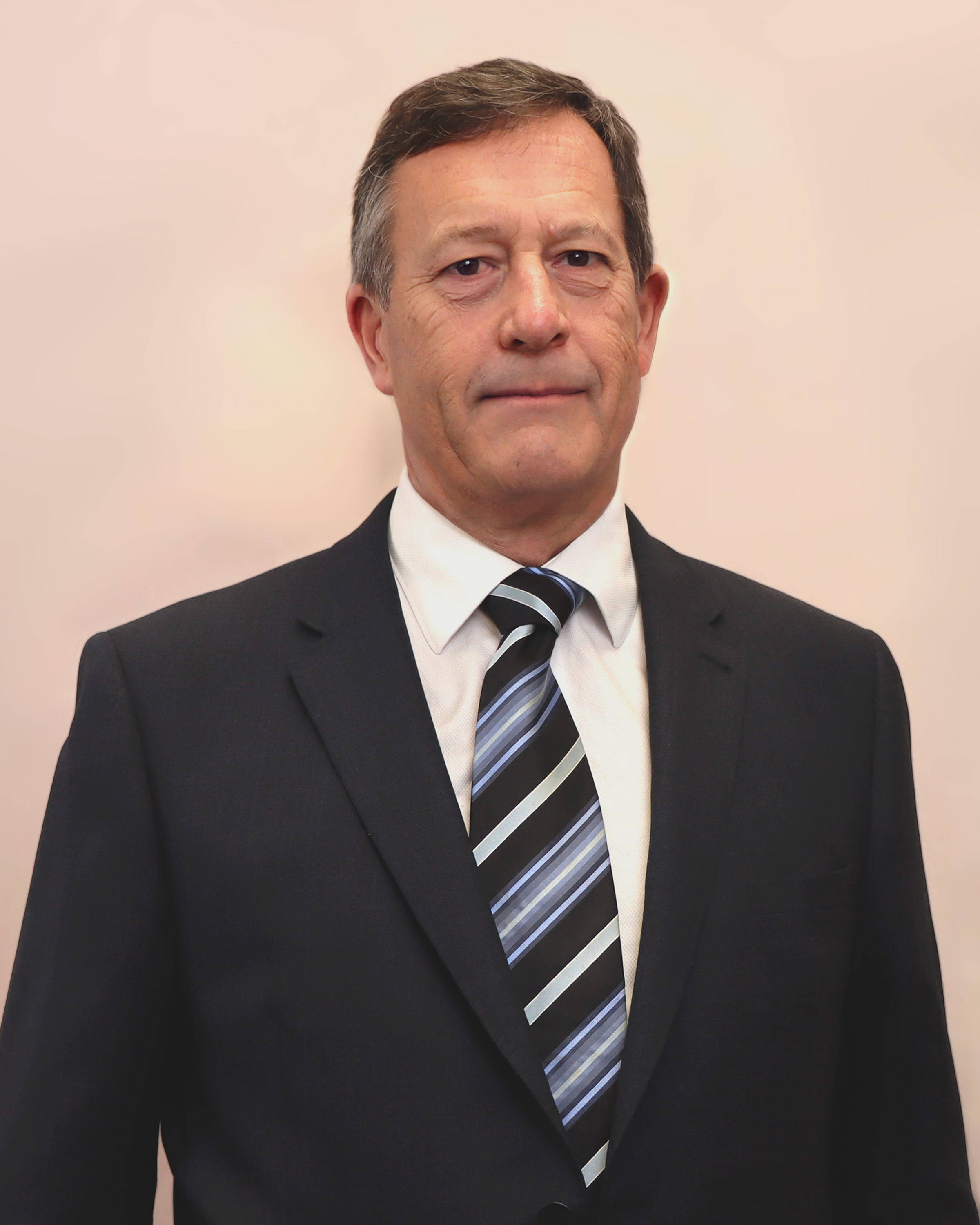 A middle-aged man wearing a black suit, white dress shirt, and striped tie standing against a plain light-colored background.