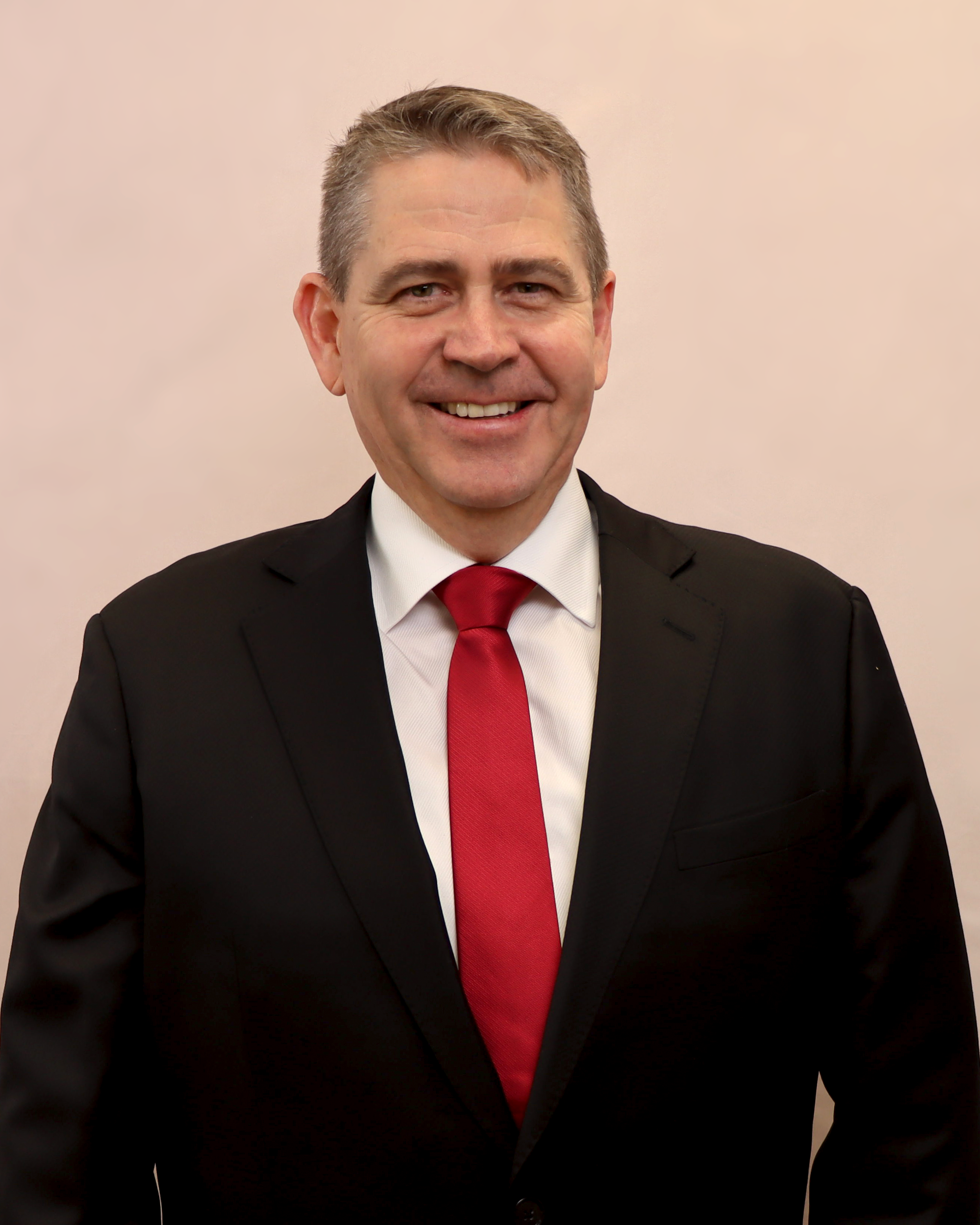 A smiling man in a black suit, white shirt, and red tie standing against a plain background.