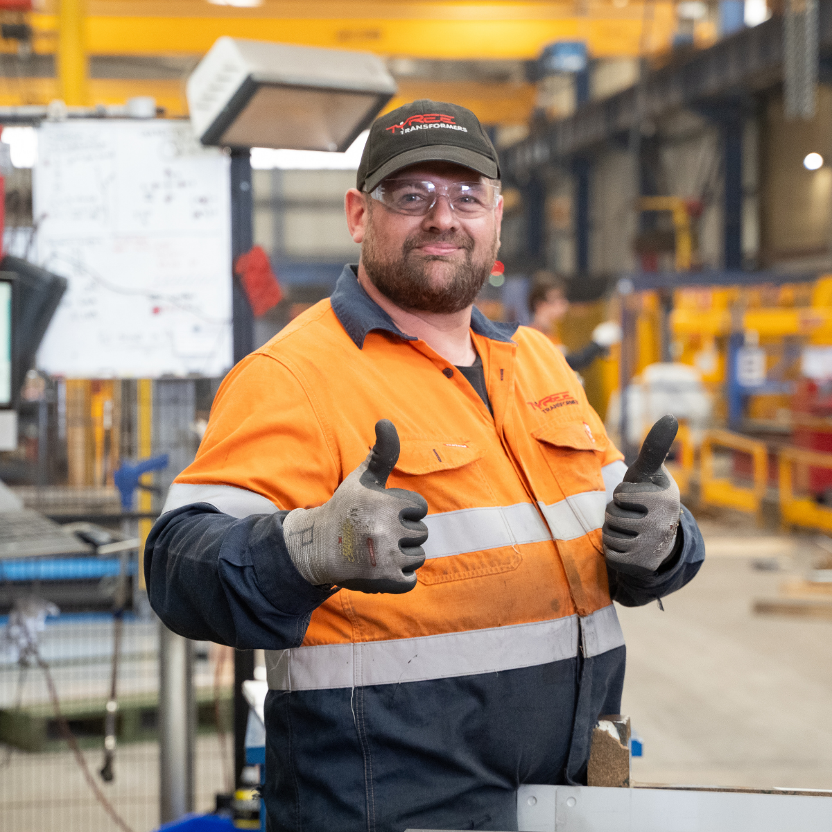 A smiling man wearing safety glasses, a black cap, an orange safety vest with reflective stripes, and work gloves, giving a thumbs-up inside an industrial warehouse or factory setting.