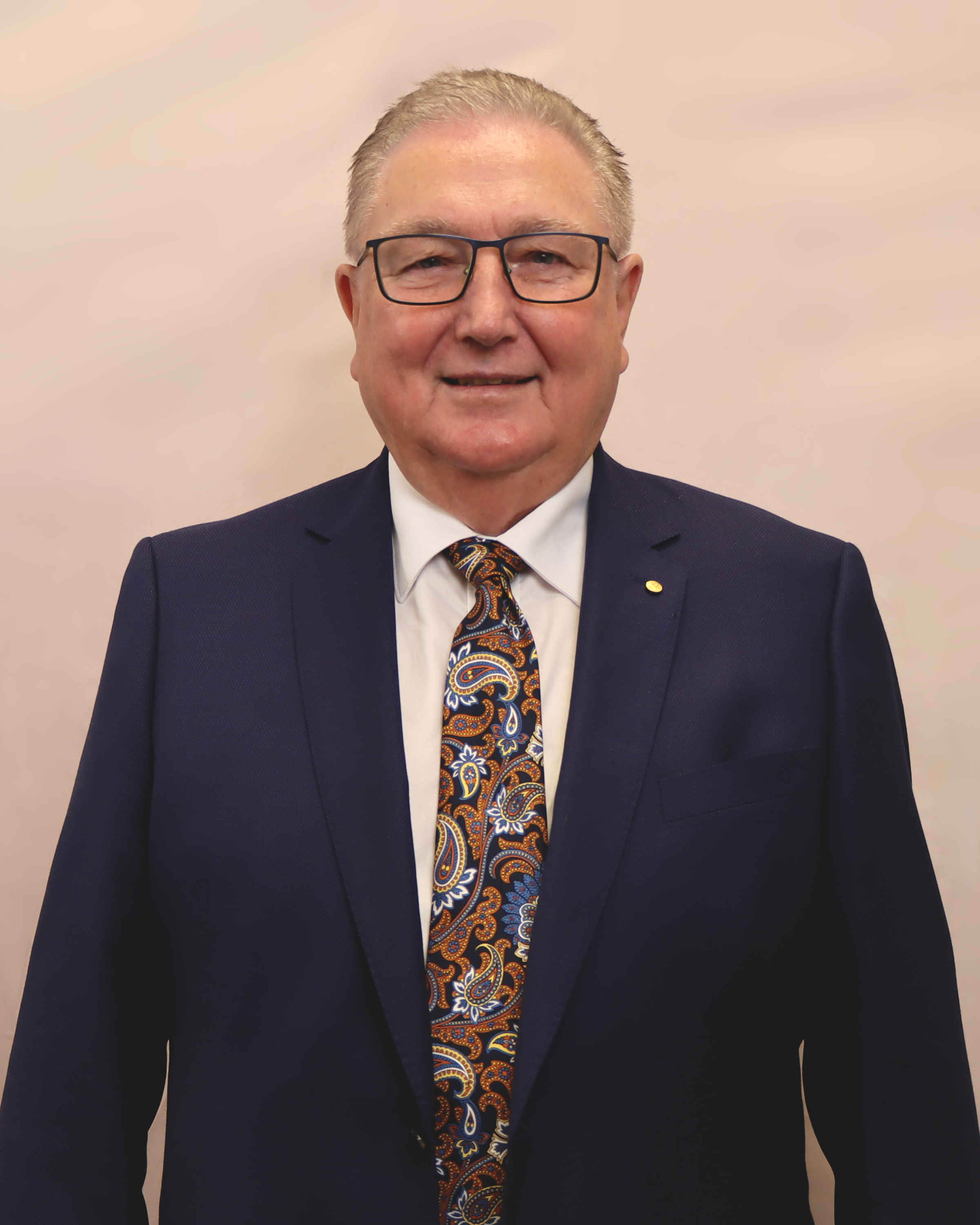 Portrait of a man in a navy suit, white shirt, and a paisley tie, wearing glasses, smiling against a neutral background.