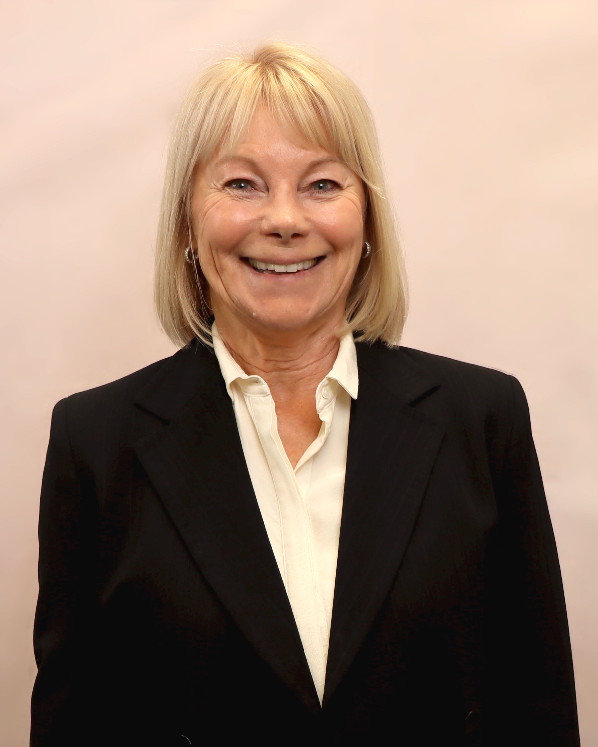 A smiling woman with blonde hair in a black blazer and white shirt posing against a plain background.