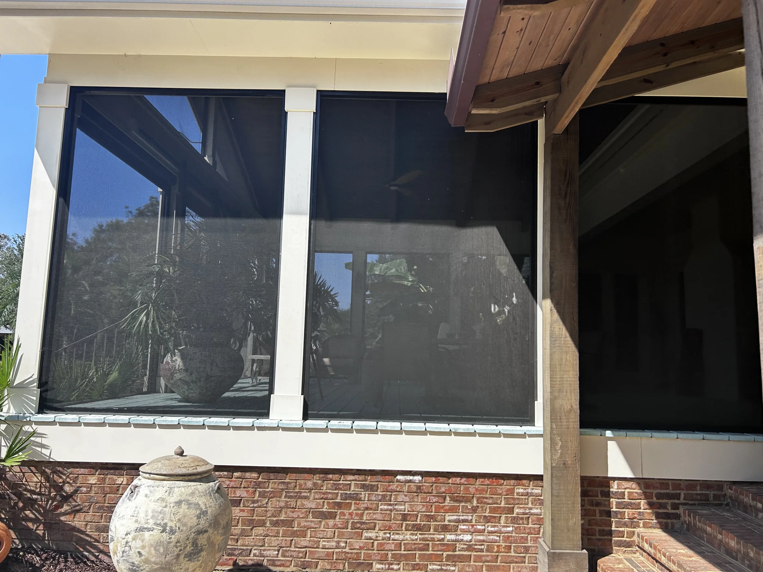 A house with large glass windows and a brick foundation, partially shaded by a wooden structure and surrounded by plants.