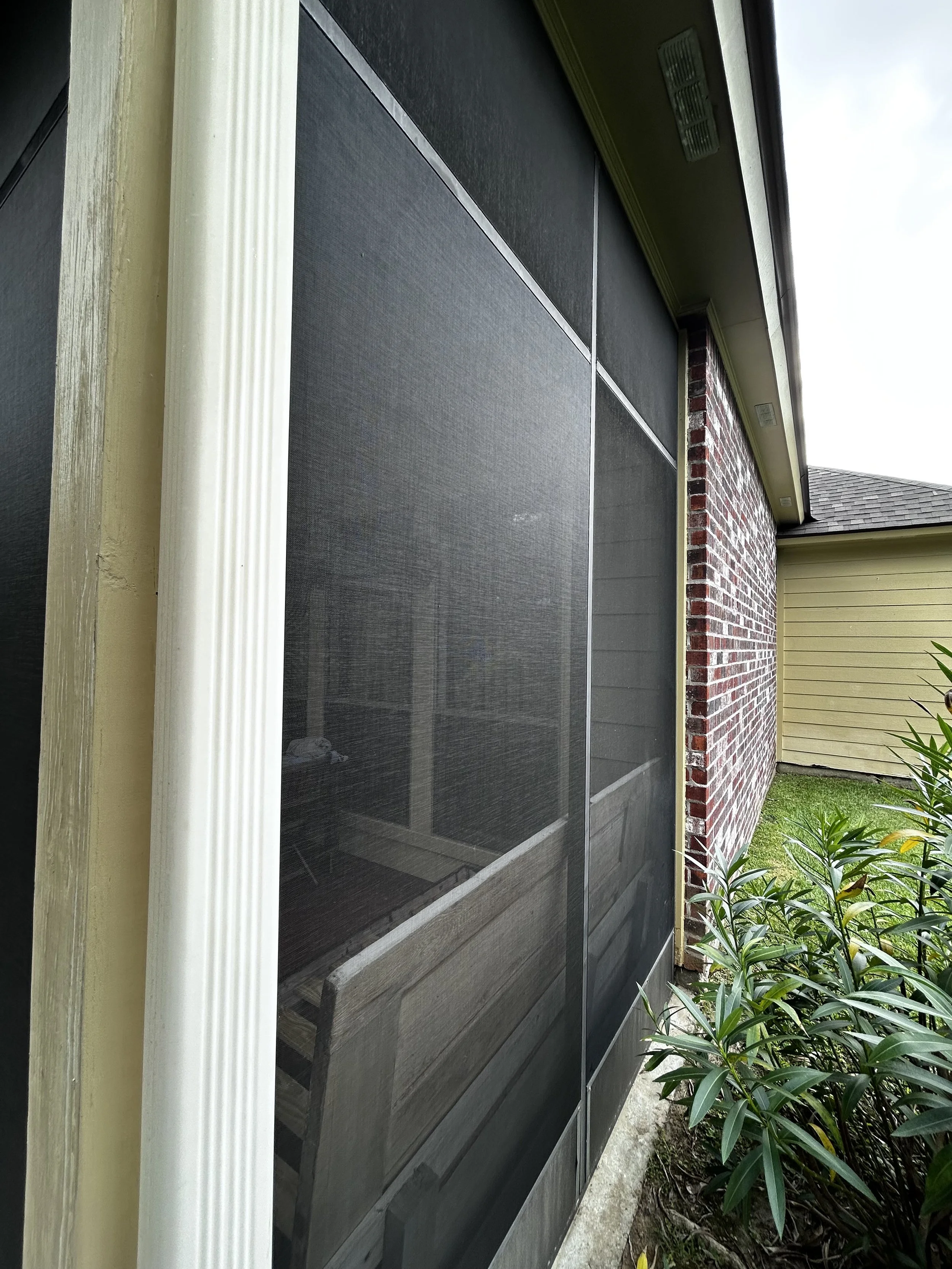 Side view of a screened porch with black mesh screens 20x20, Solid Kick Plate, and a brick wall on one side. There are green bushes and grass outside.