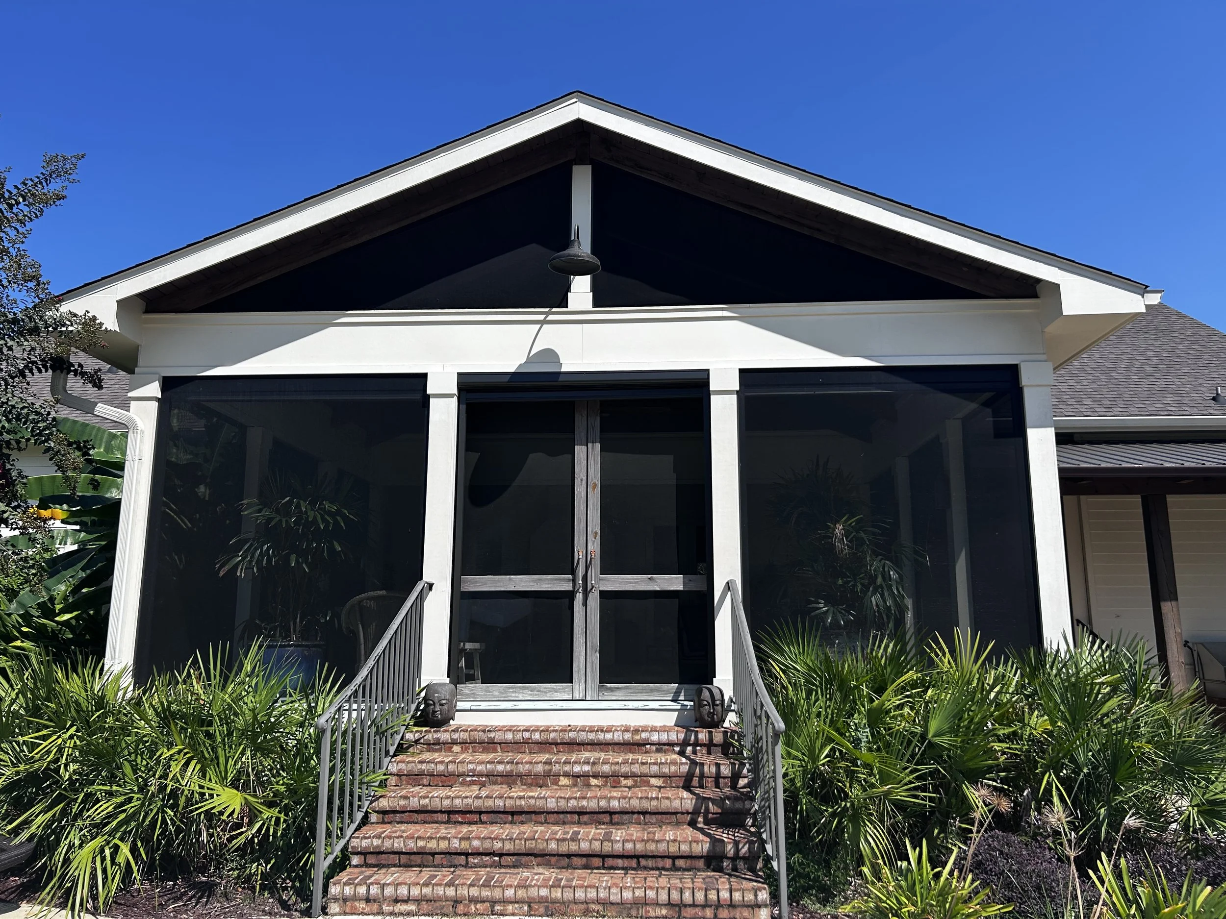 Front view of a house with a screened porch, brick steps, railing, plants, and a gable roof against a blue sky.
