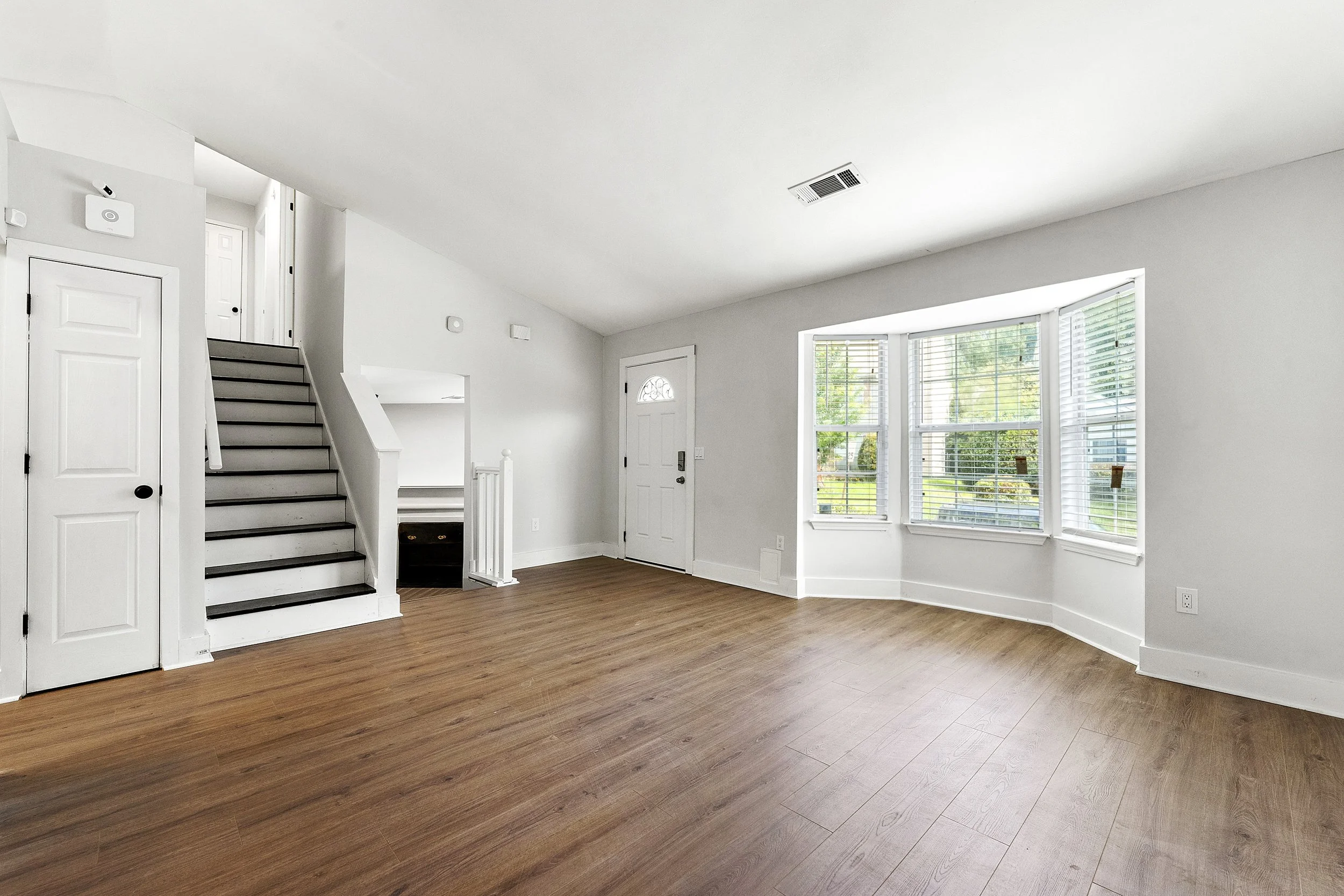 Empty living room with white walls, hardwood floors, a staircase, and large bay windows with blinds overlooking a green yard.