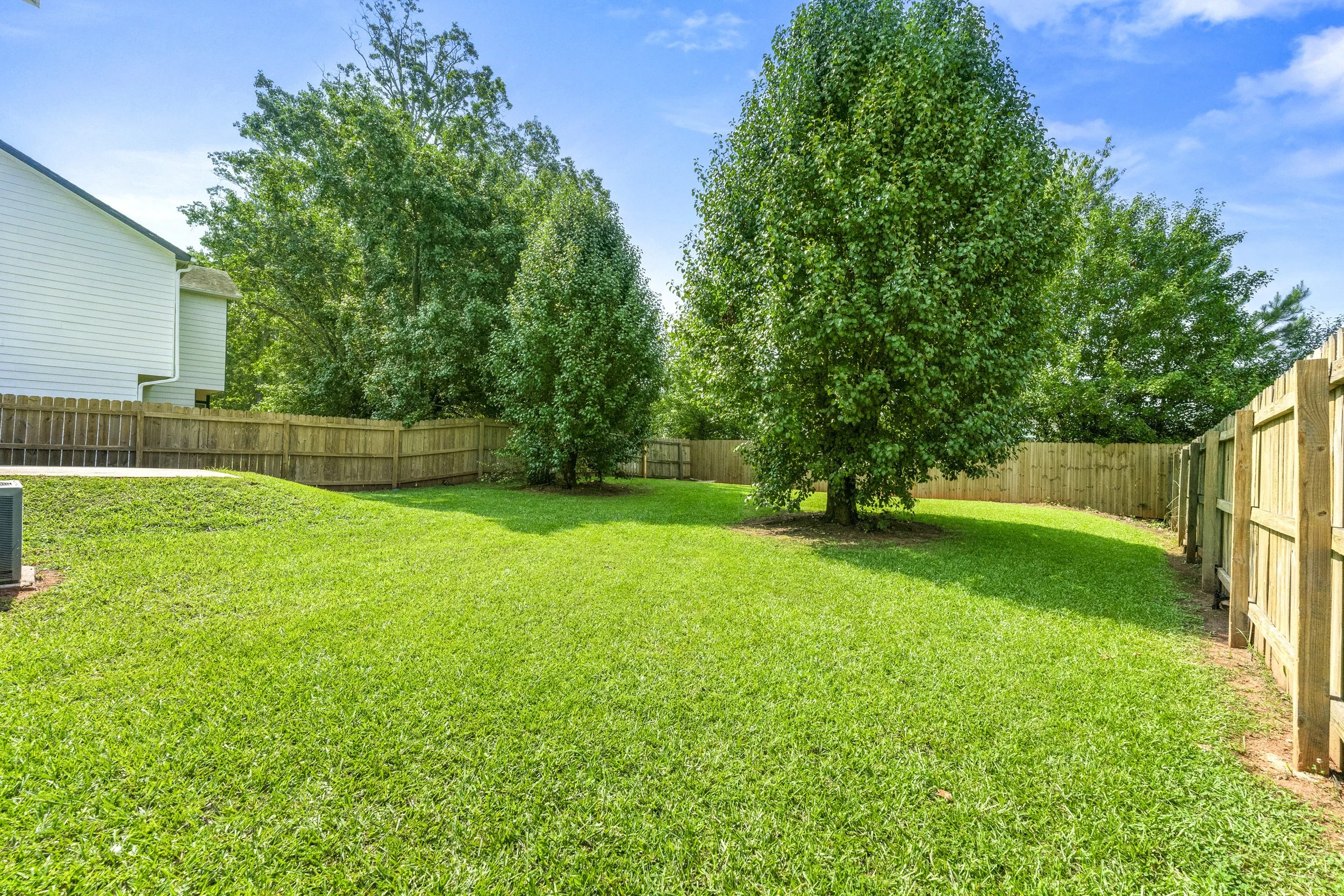 Backyard with green grass, two large trees, wooden fence, and part of a white house with outside air conditioning unit
