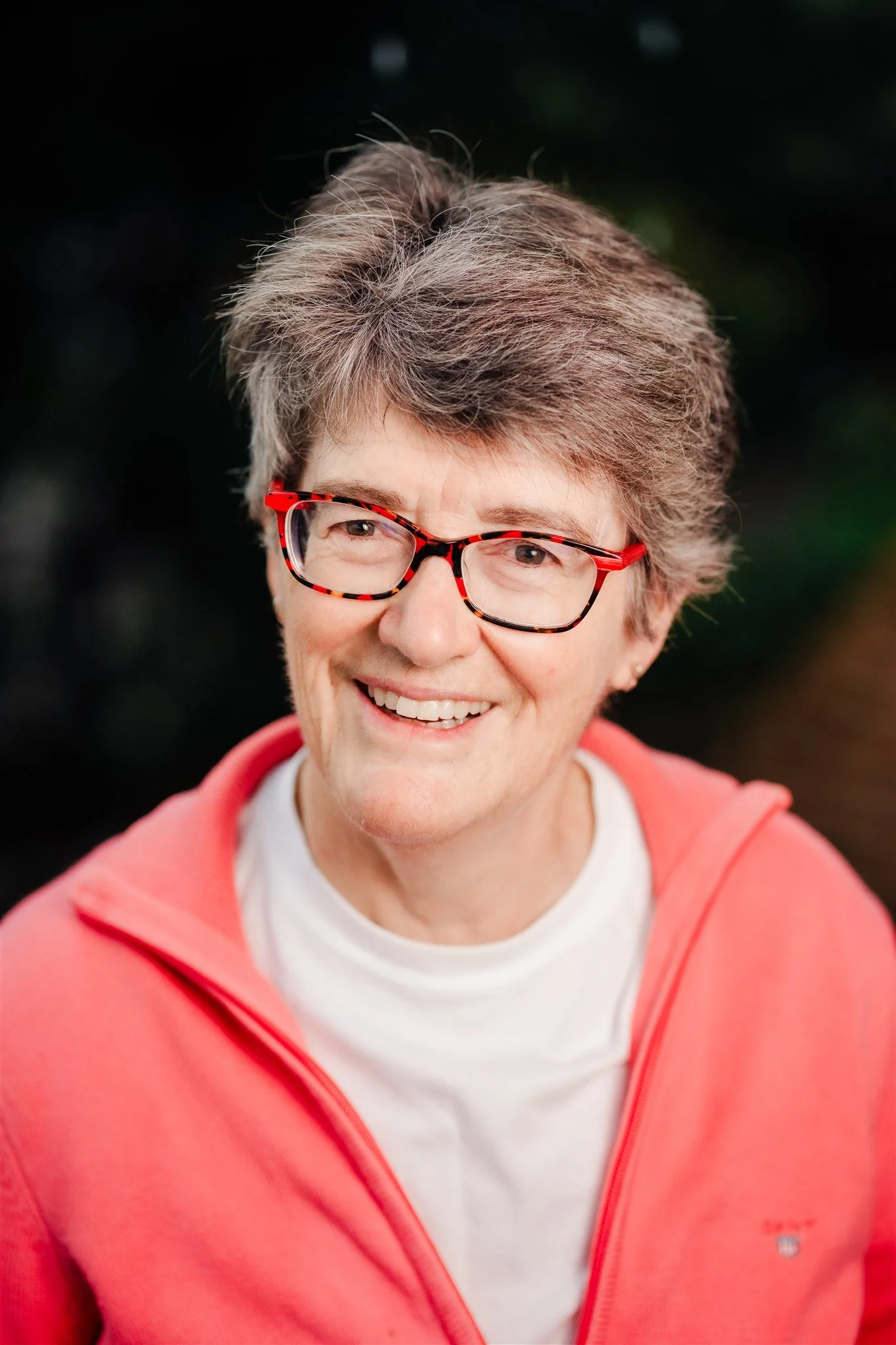 Di Morgan is smiling to camera. She has cropped grey hair, red glasses and dangly earrings. She wears a white t-shirt and black cardigan. She is standing in the library at Mary Andrews College.