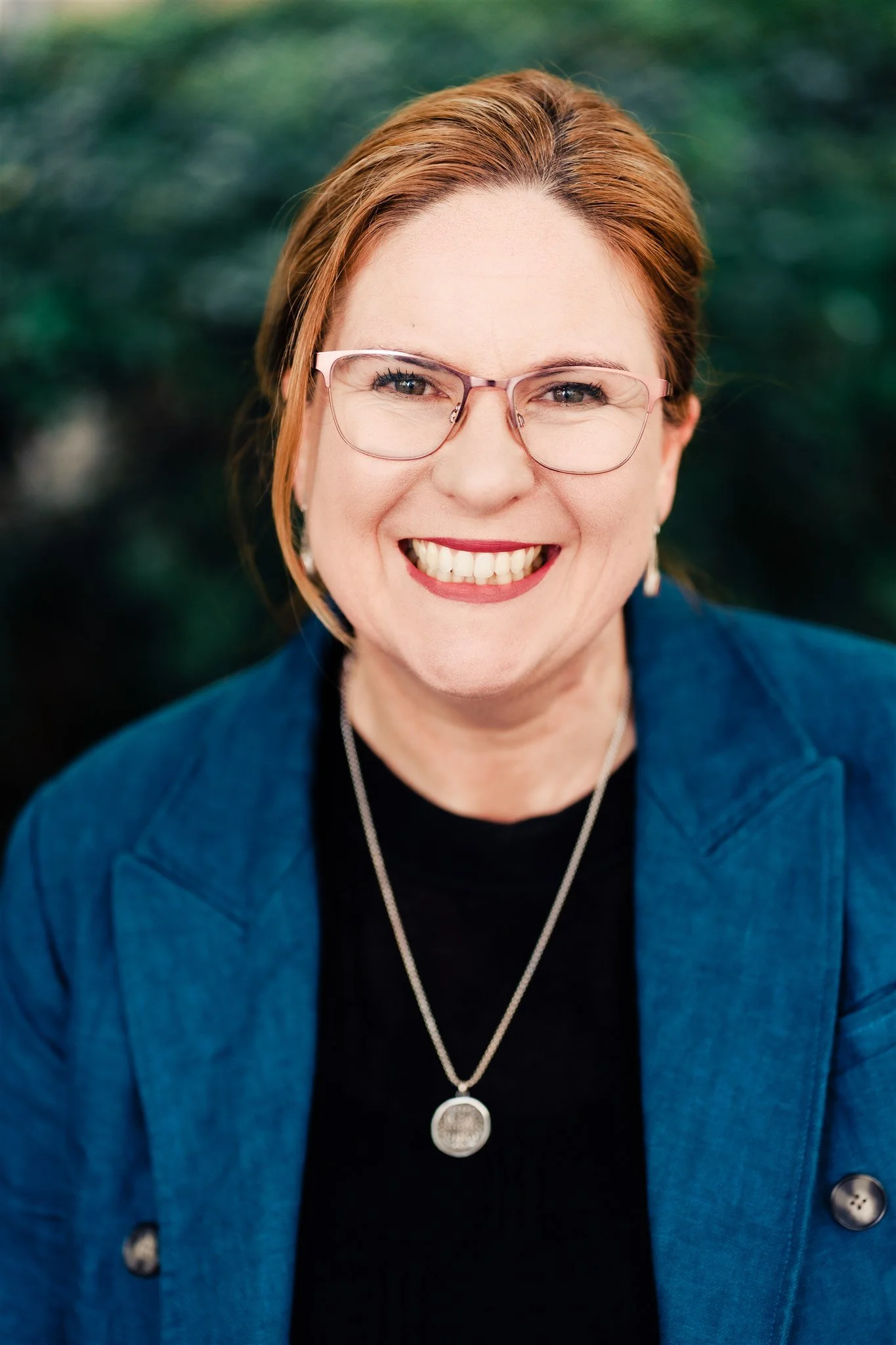Dr Nicole Starling has brown hair, styled in a bob and is wearing a link and brown dress. The photo shows her smiling to camera, standing in the library at Mary Andrews College