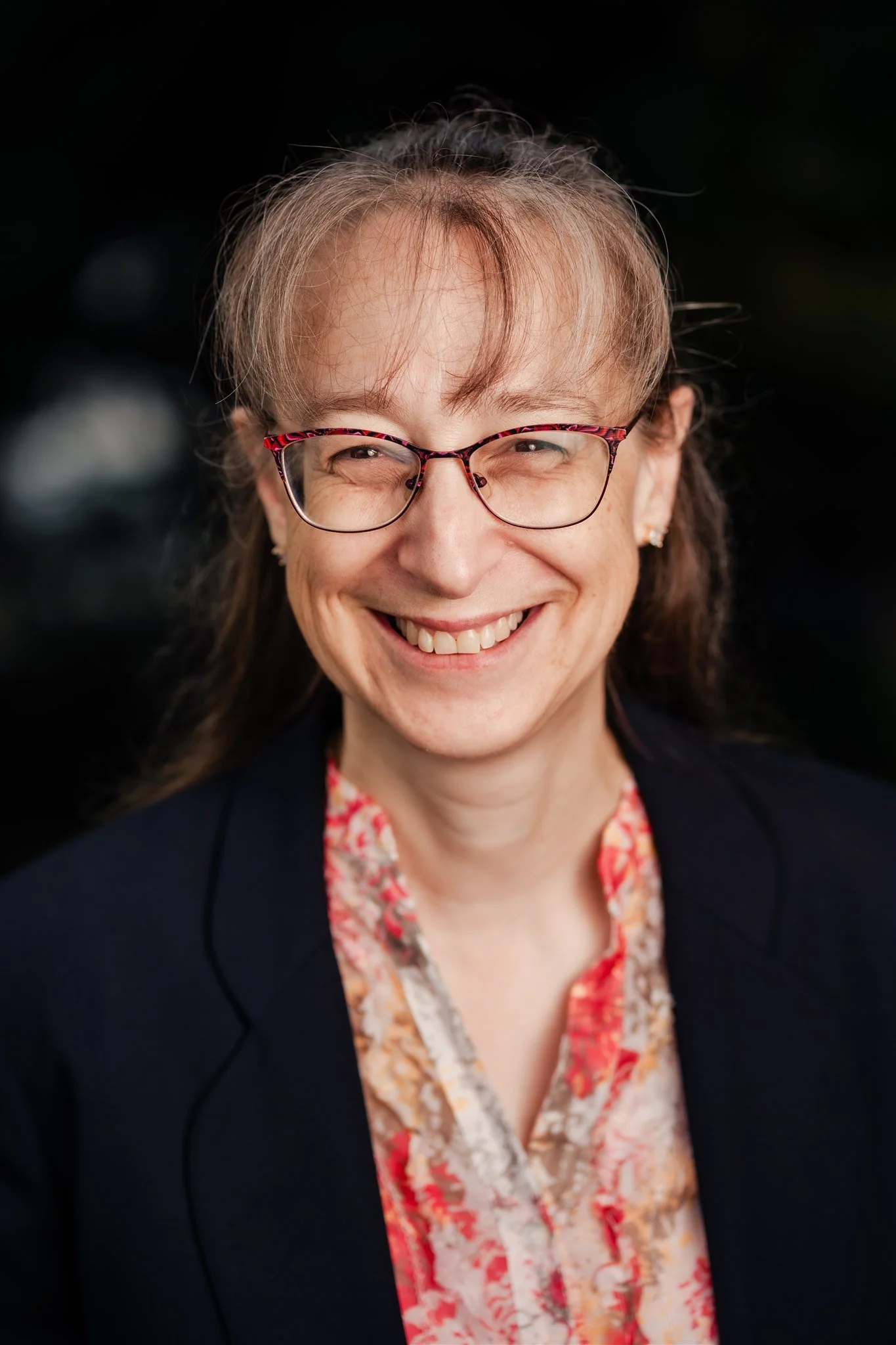 Sarah has long light brown hair, with a fringe, tied back off her face. She is wearing a red dress and taupe cardigan. She is smiling at the camera and wears glasses.