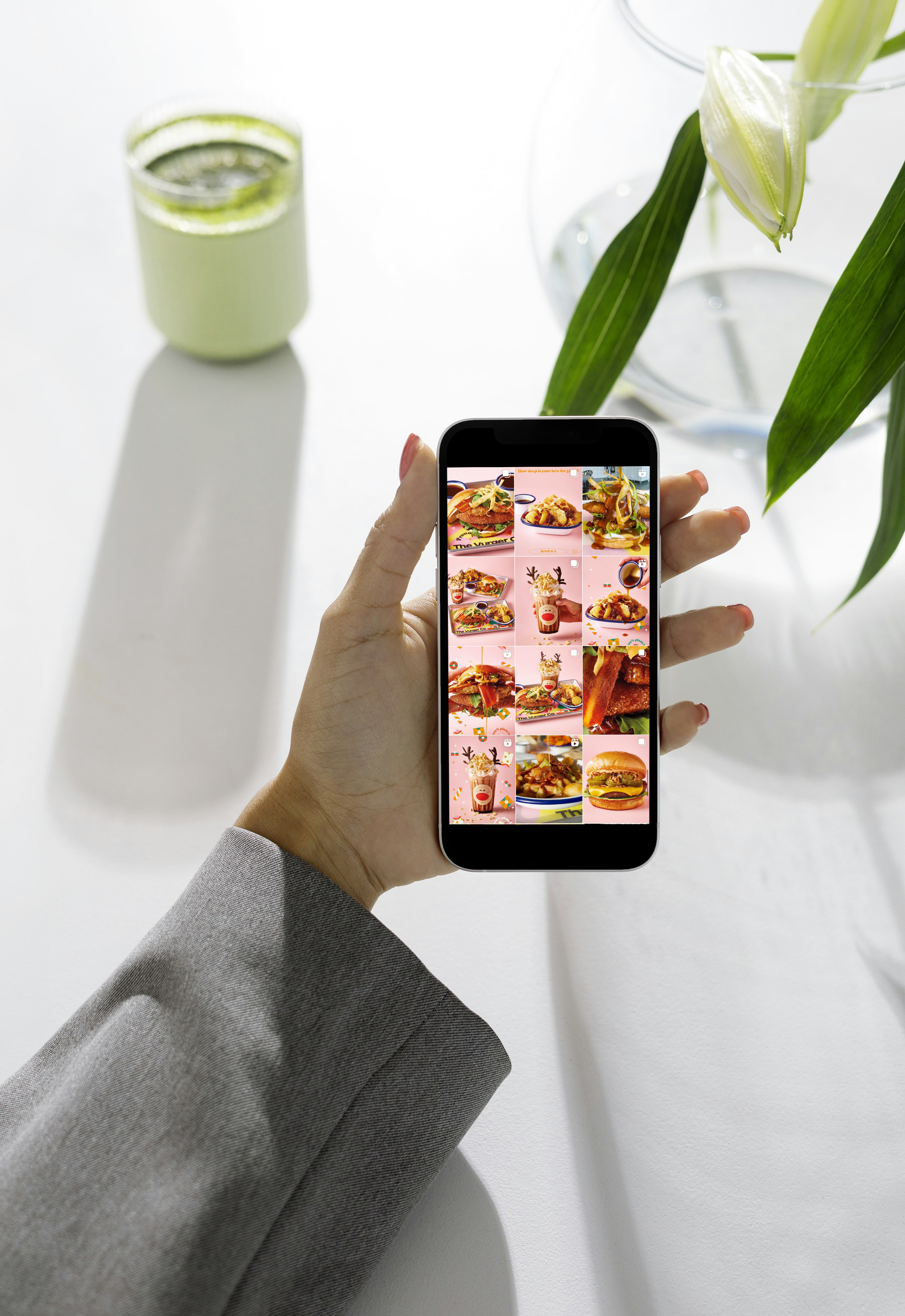 A person's hand holds a smartphone displaying a grid of food images, with a white table, a glass jar of flowers, and a small jar of cucumber slices in the background.