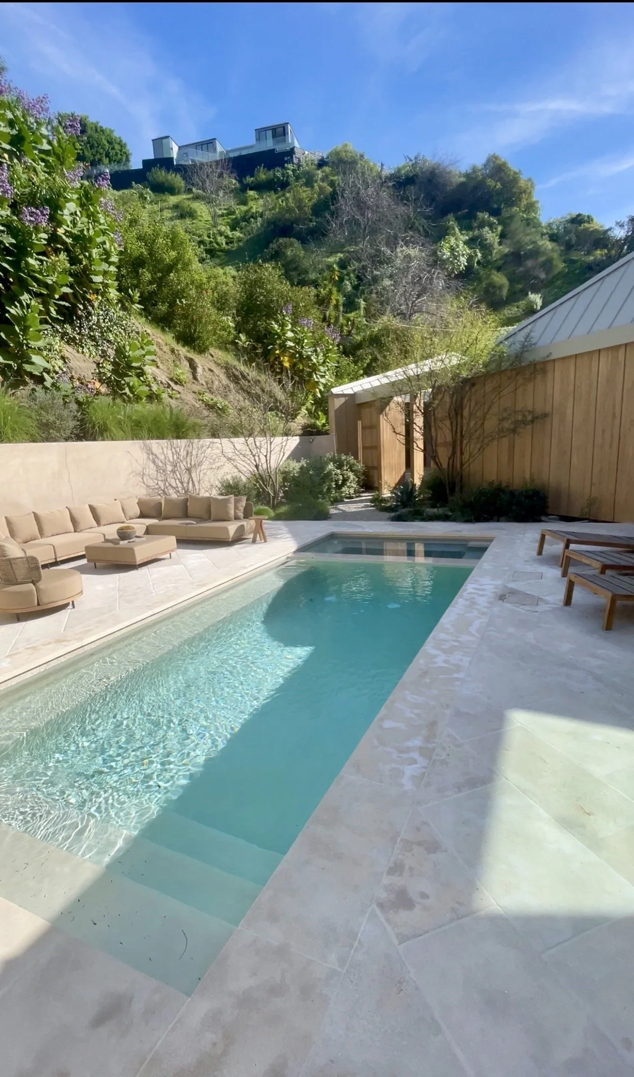 Luxurious backyard with a rectangular swimming pool, beige outdoor sofa, and wooden lounge chairs, surrounded by greenery and hillside homes in the background on a sunny day.