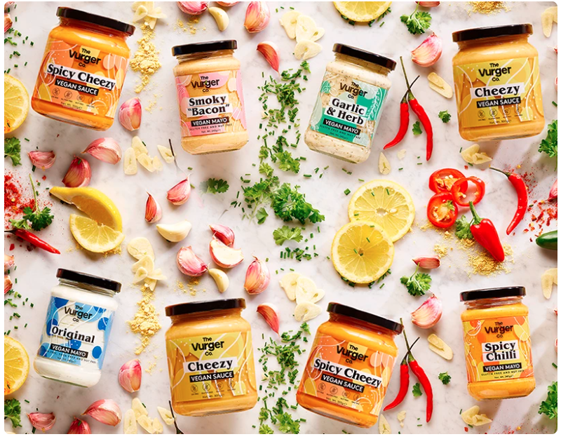 A flat lay display of eight jars of vegan sauce with various flavors, surrounded by garlic cloves, lemon slices, red chili peppers, chopped herbs, and garlic pieces on a white surface.