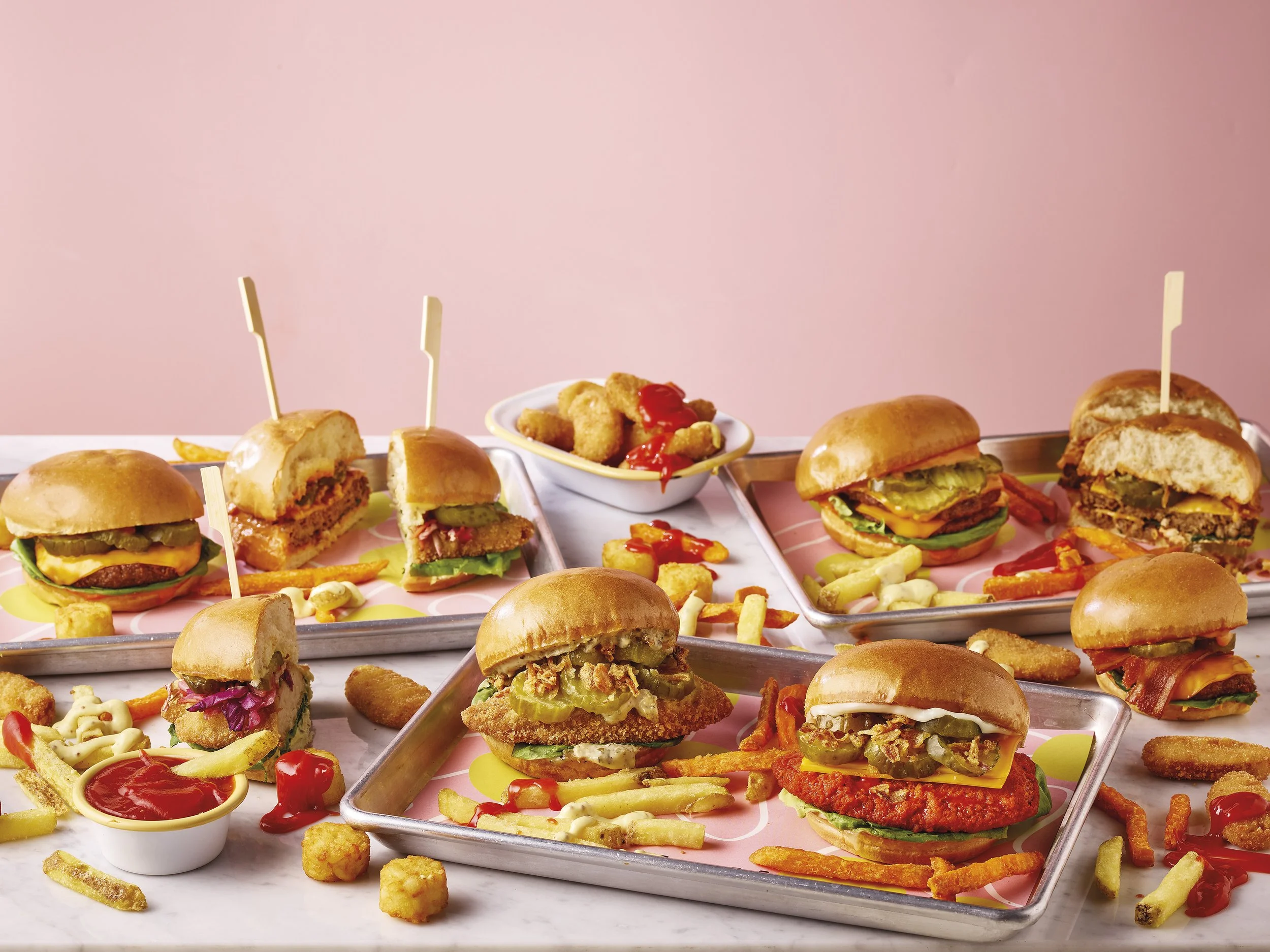 An assortment of various burgers, chicken nuggets, French fries, onion rings, and dipping sauces on metal trays and bowls against a pink background.