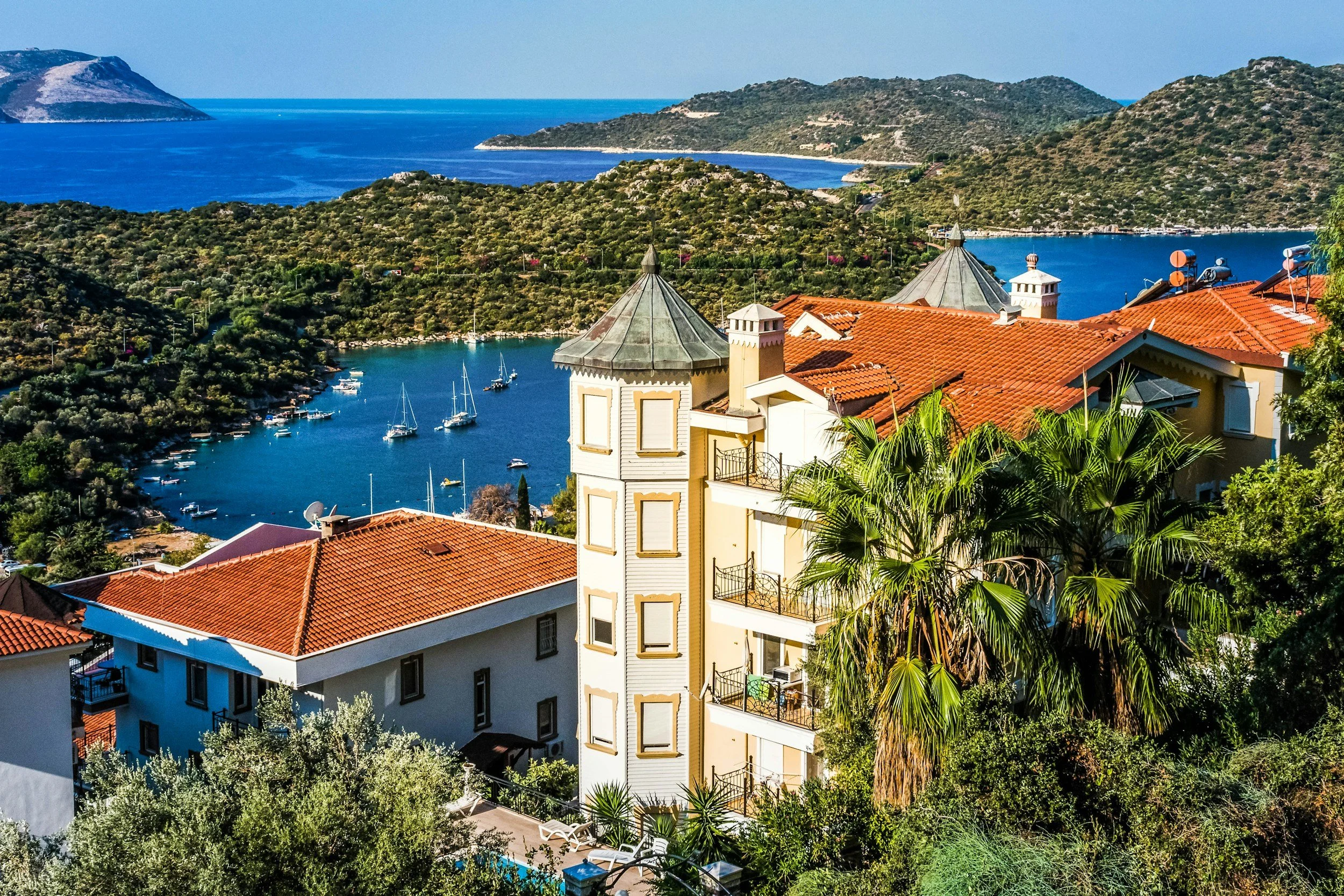 Välimerellinen rannikko, pehmeästi kalkkikoristeinen rakennus ja palmupuita, taustalla vuoret ja meri