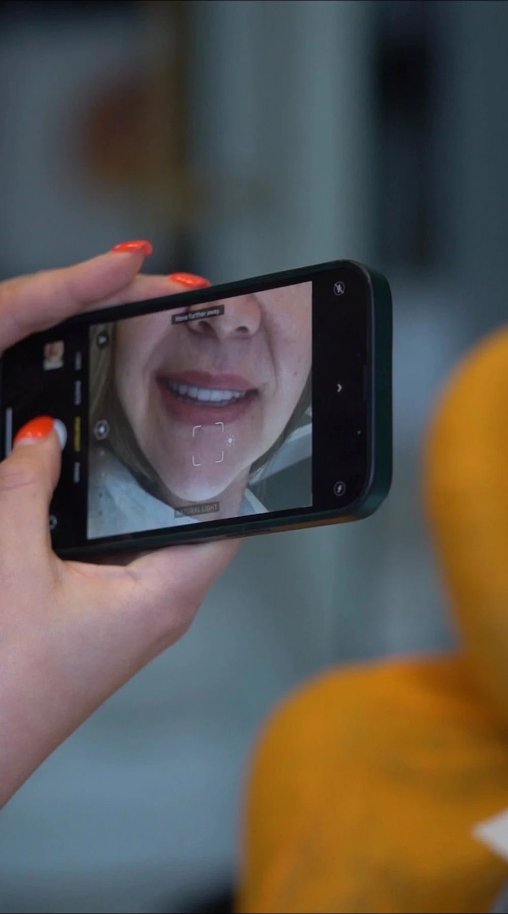 Henkilö ottaa selfie-kuvaa älypuhelimella, näytöllä näkyy naisen kasvot ja hymy, taustalla näkyy oranssi.