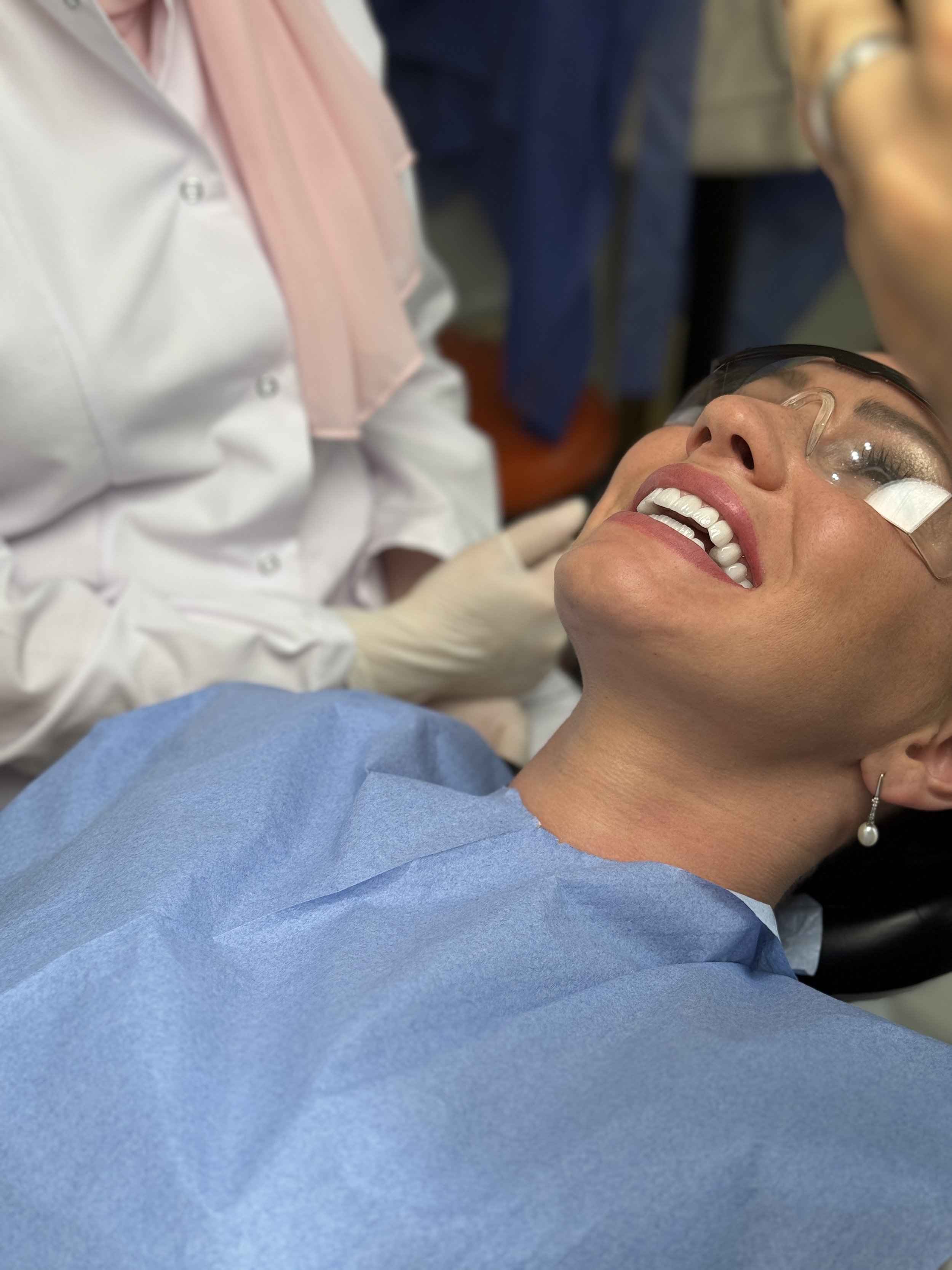 Nainen hammashoitajan hoivassa, makaa hoitopöydällä, suojalaseissa, hymyilee, hammaslääkärin ja hoitovälineiden ympäröimänä.