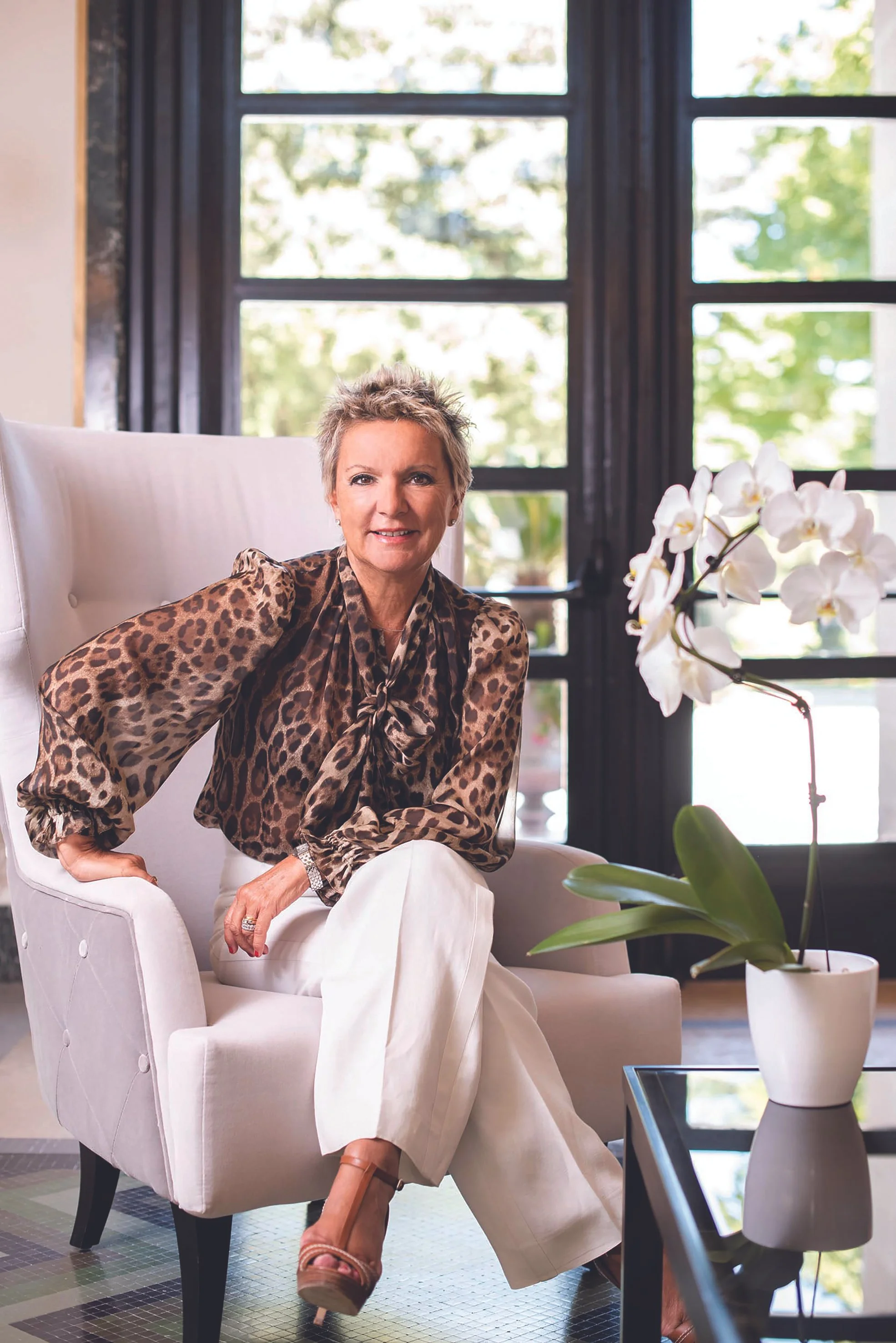 Nainen istuu valkoisessa nojatuolissa, ympärillään kukkavaasi ja suuri ikkuna taustalla, joka avautuu vihreään pihamaahan.