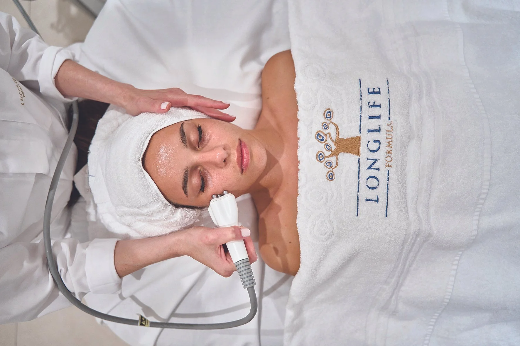 Nainen makaa hoidettavaksi, hänellä on pyyhe päässään ja yllä valkoinen kylpytakki, jossa lukee "LONGLIFE FOR KAILUE". Hoitaja pitää laitetta naisen kasvoilla ja tukee hänen päätään.