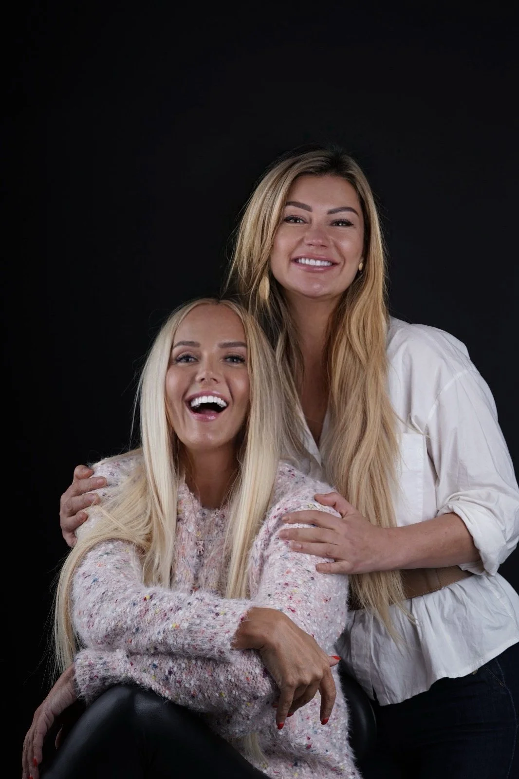 Kaksi naista hymyilee ja halailee toisiaan mustassa taustassa.