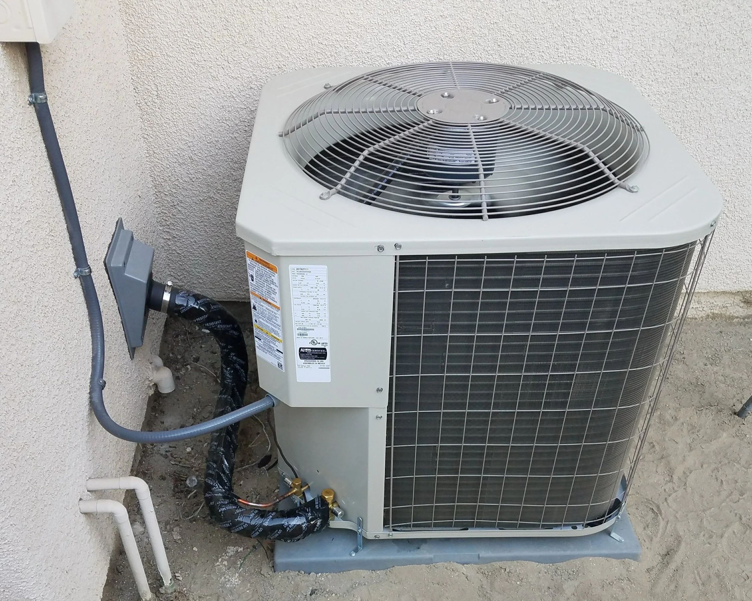 Outdoor air conditioning unit installed on concrete slab next to a beige stucco wall with electrical wiring and pipes.
