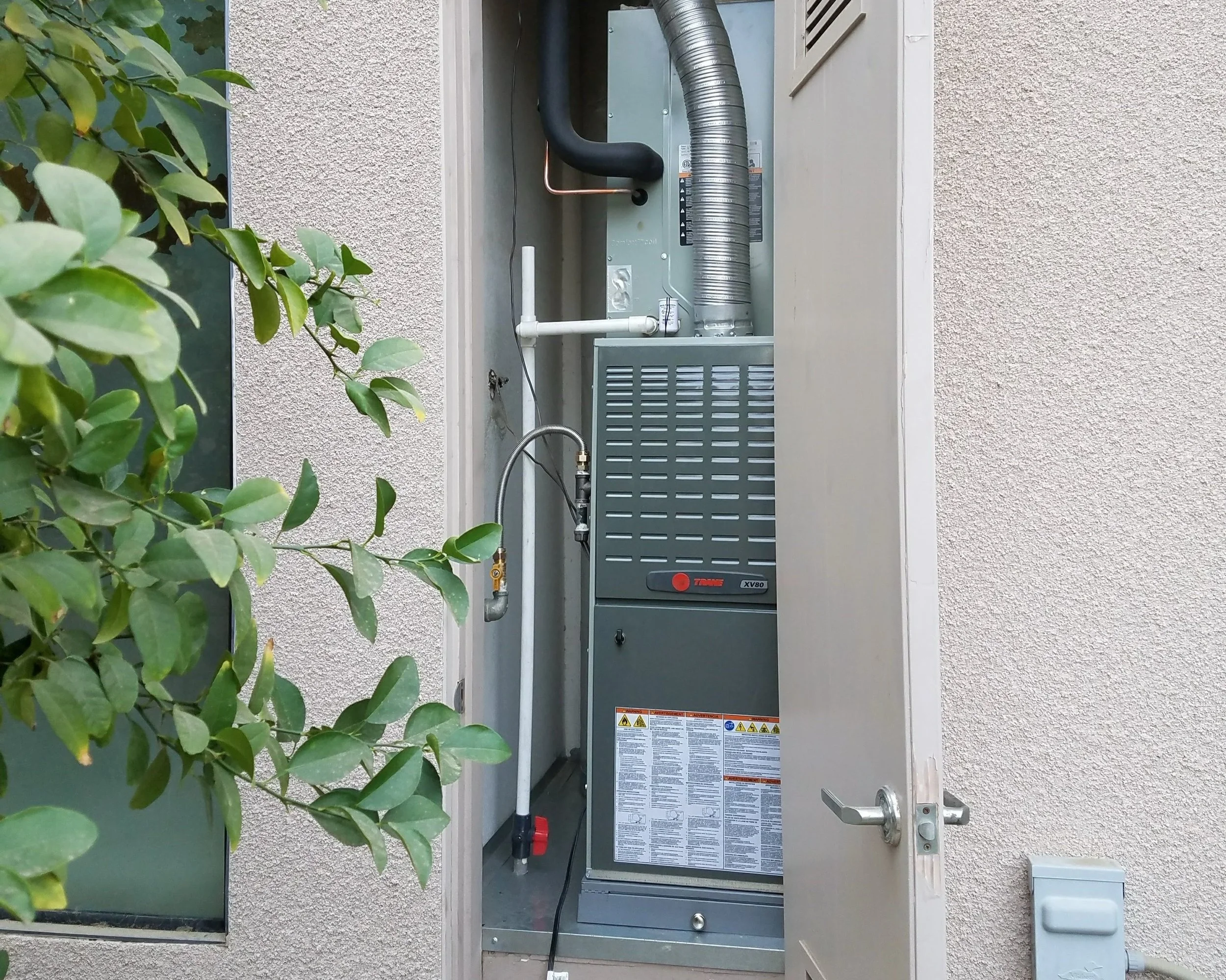 An electrical or HVAC unit installed in an outdoor utility closet, partially opened door, on a beige textured wall, with green leaves from a plant visible on the left.