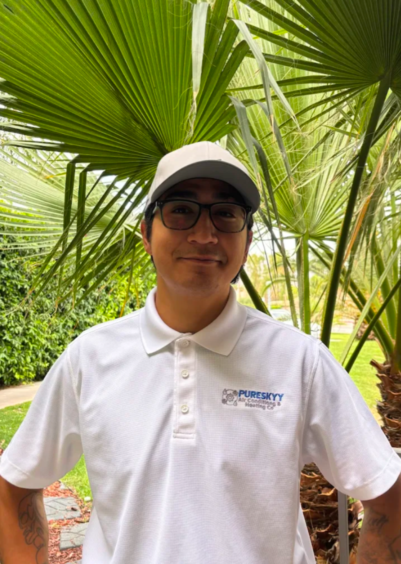 Smiling man, company founder, wearing glasses, a white cap, and a white polo shirt with the logo of PURESKYY Air Conditioning Heating Co. Stands outdoors in front of large green palm leaves and lush garden greenery.