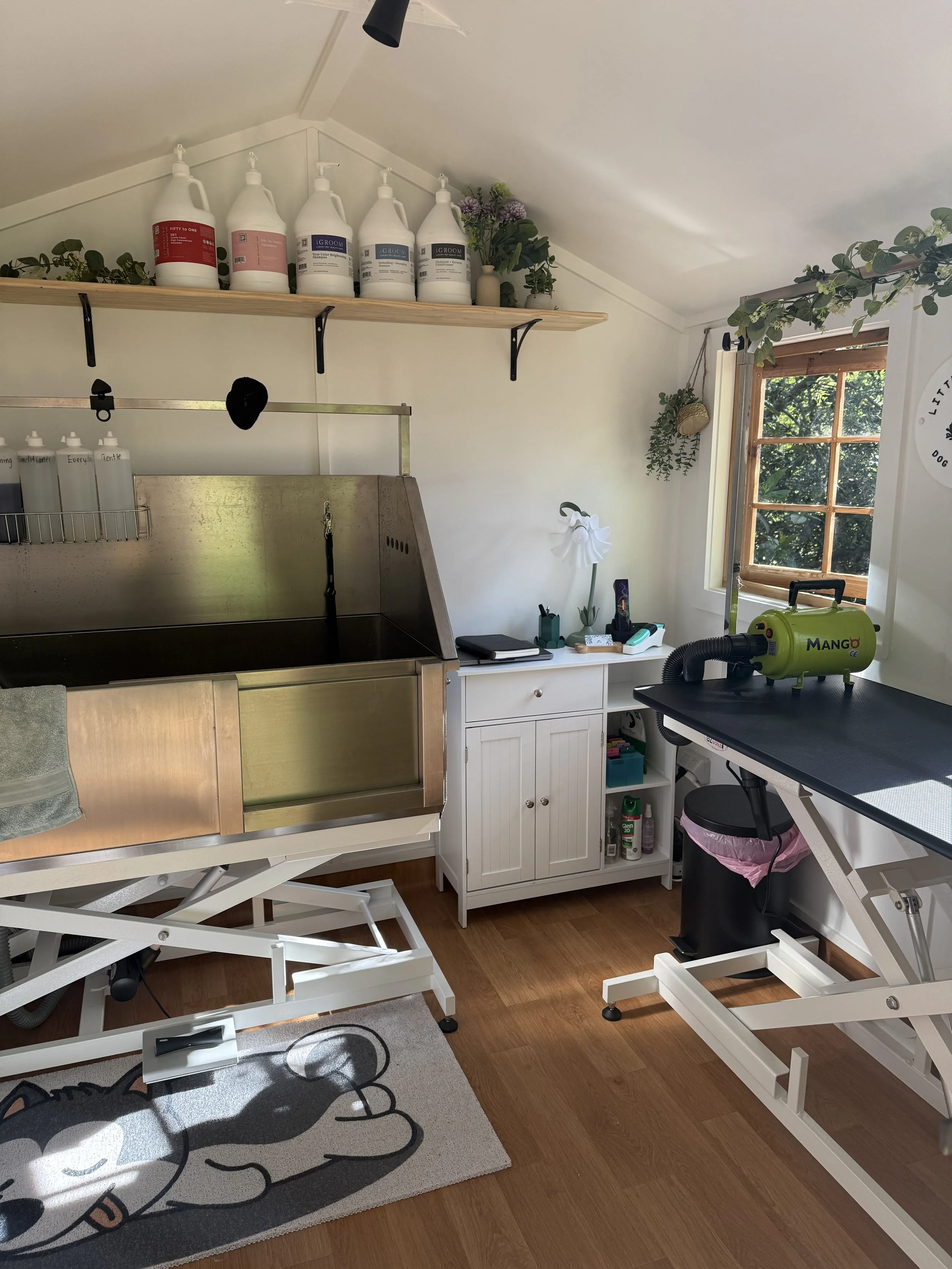 A grooming room with a grooming table, a sink, and supplies. There are cleaning products on a wooden shelf, a window with greenery outside, and grooming tools on a white cabinet. A dog grooming vacuum is on a black table, and a trash can is nearby.