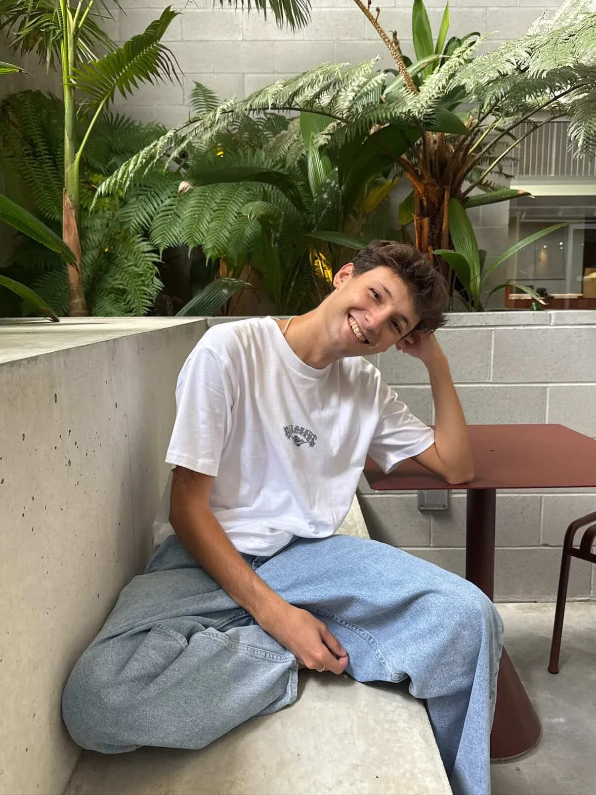 A young man sitting on a concrete bench, smiling, wearing a white t-shirt and gray pants, with a background of lush green plants and a gray wall.