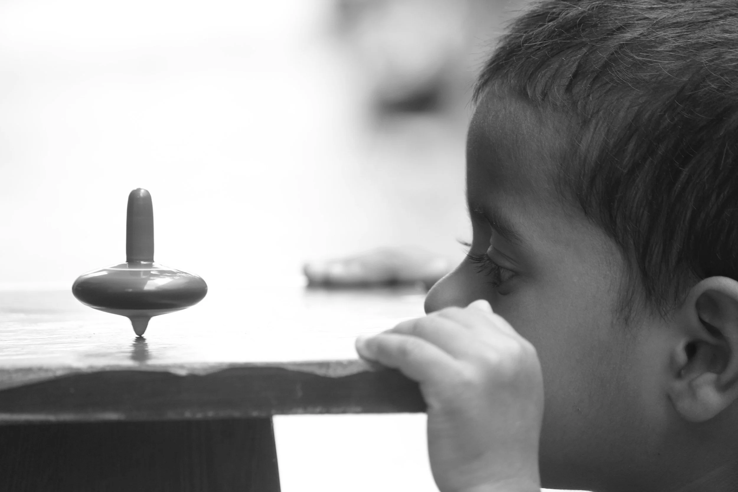 A young boy with short hair watches a spinning top on a table in black and white.