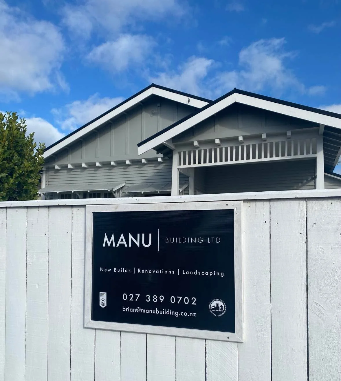 A white fence with a sign for MANU Building Ltd, showing contact information and services, in front of a modern house with gray siding and white trim against a blue sky with clouds.