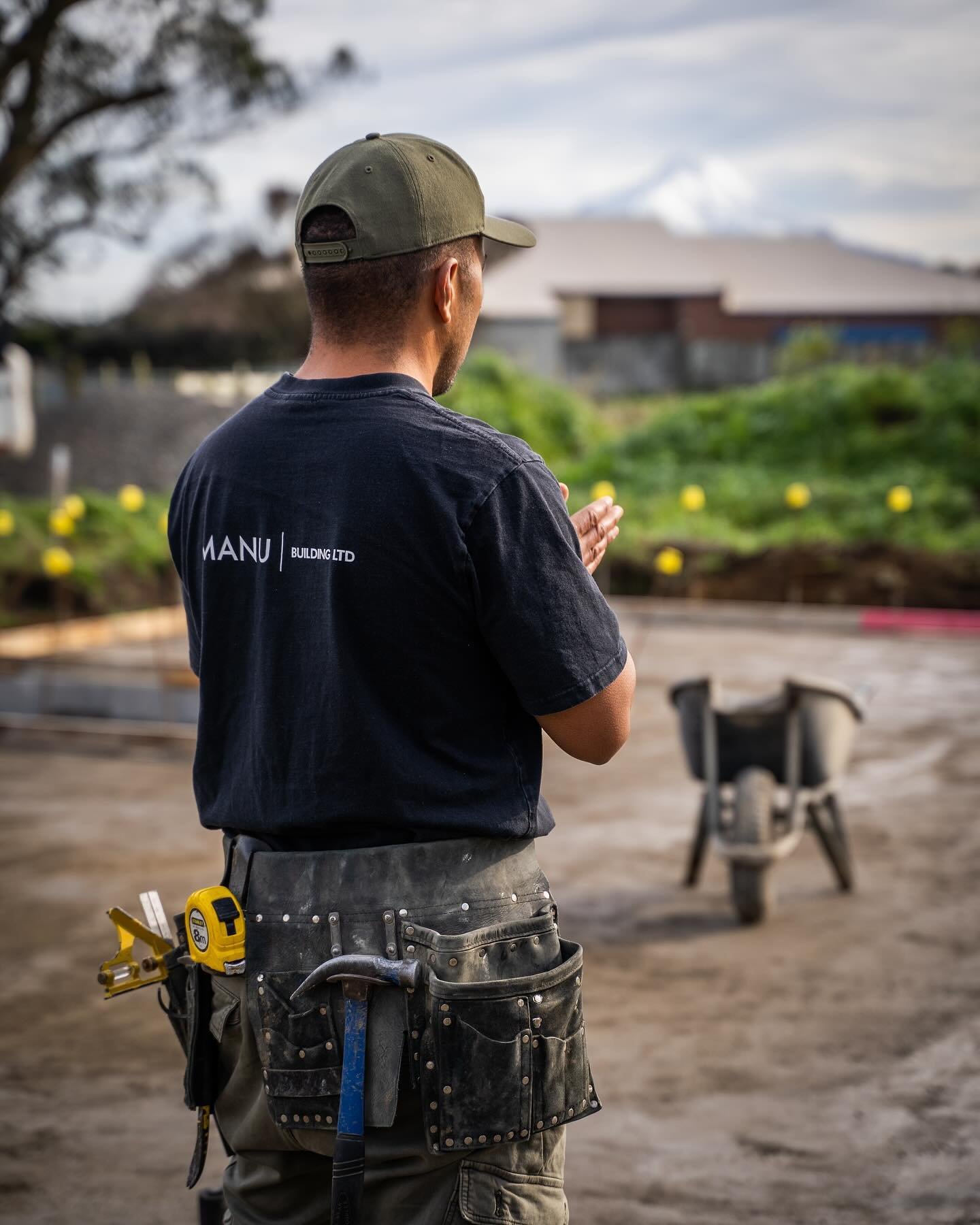 Construction worker standing with back to camera, hands together, at a construction site with a wheelbarrow and greenery in the background.
