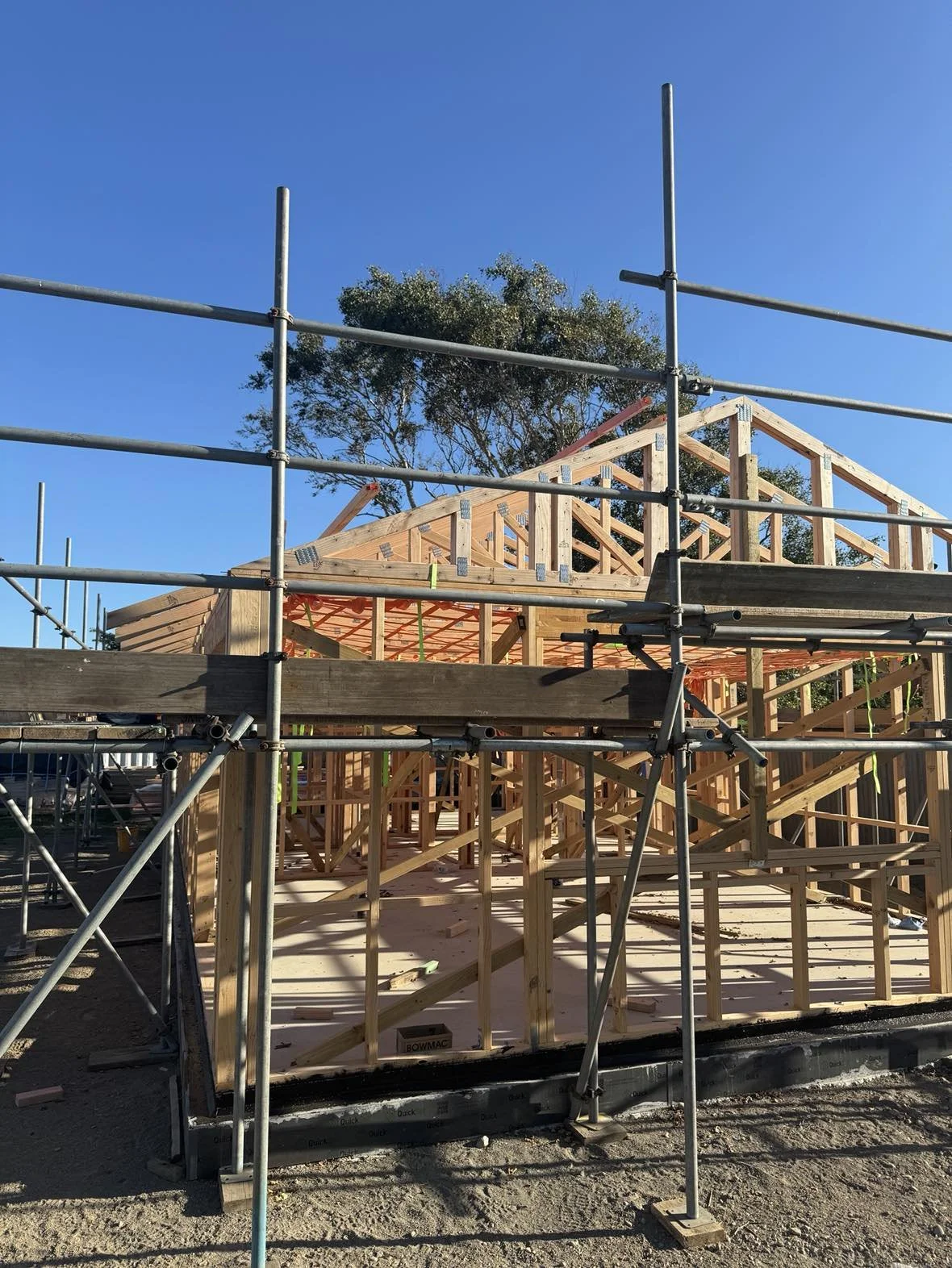 Wooden house frame under construction with scaffolding around it, a clear blue sky, and a tree in the background.