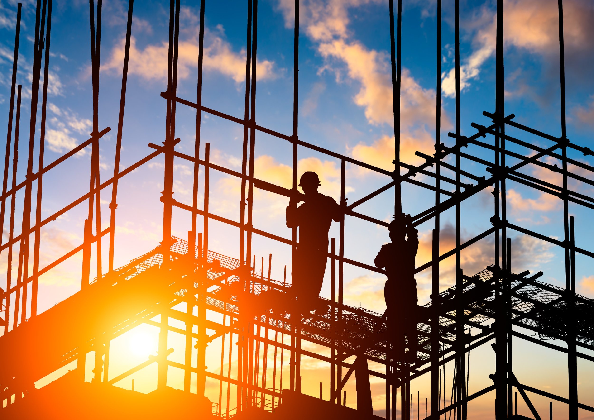 Silhouettes of construction workers on scaffolding against a sunset sky with clouds.