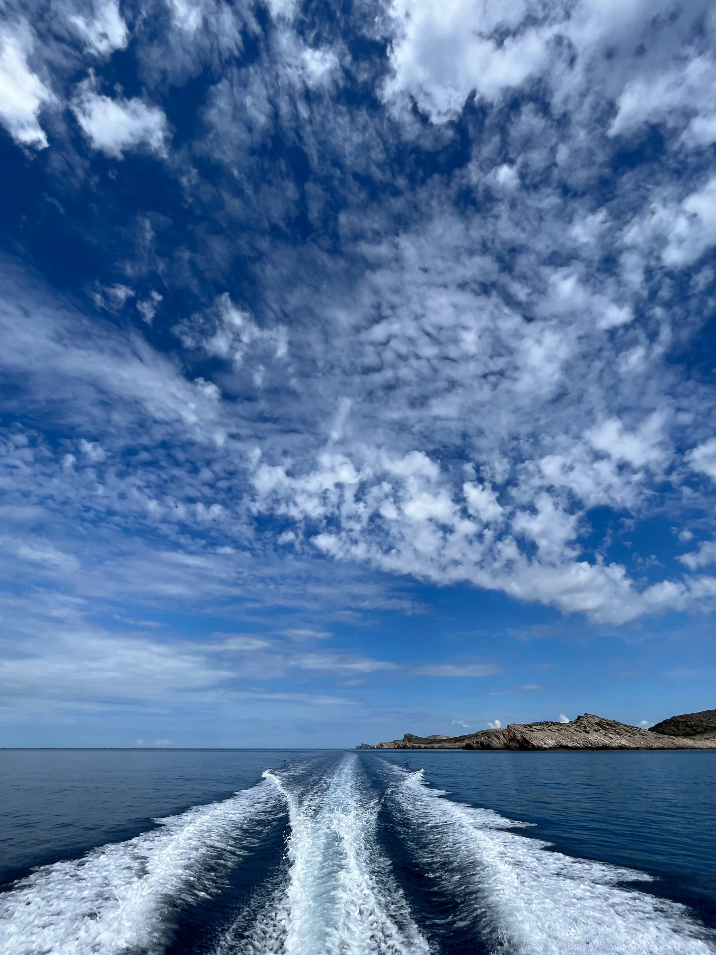 View from a boat on the water with a trail of white foam behind it, a distant rocky Kornati shoreline, and a blue sky with scattered clouds.
