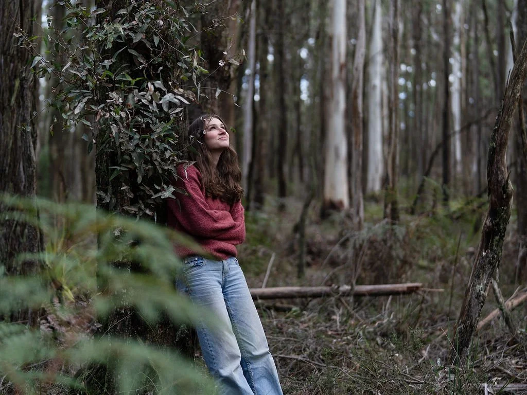 young lady in jeans and jumper in forest leaning on tree looking up