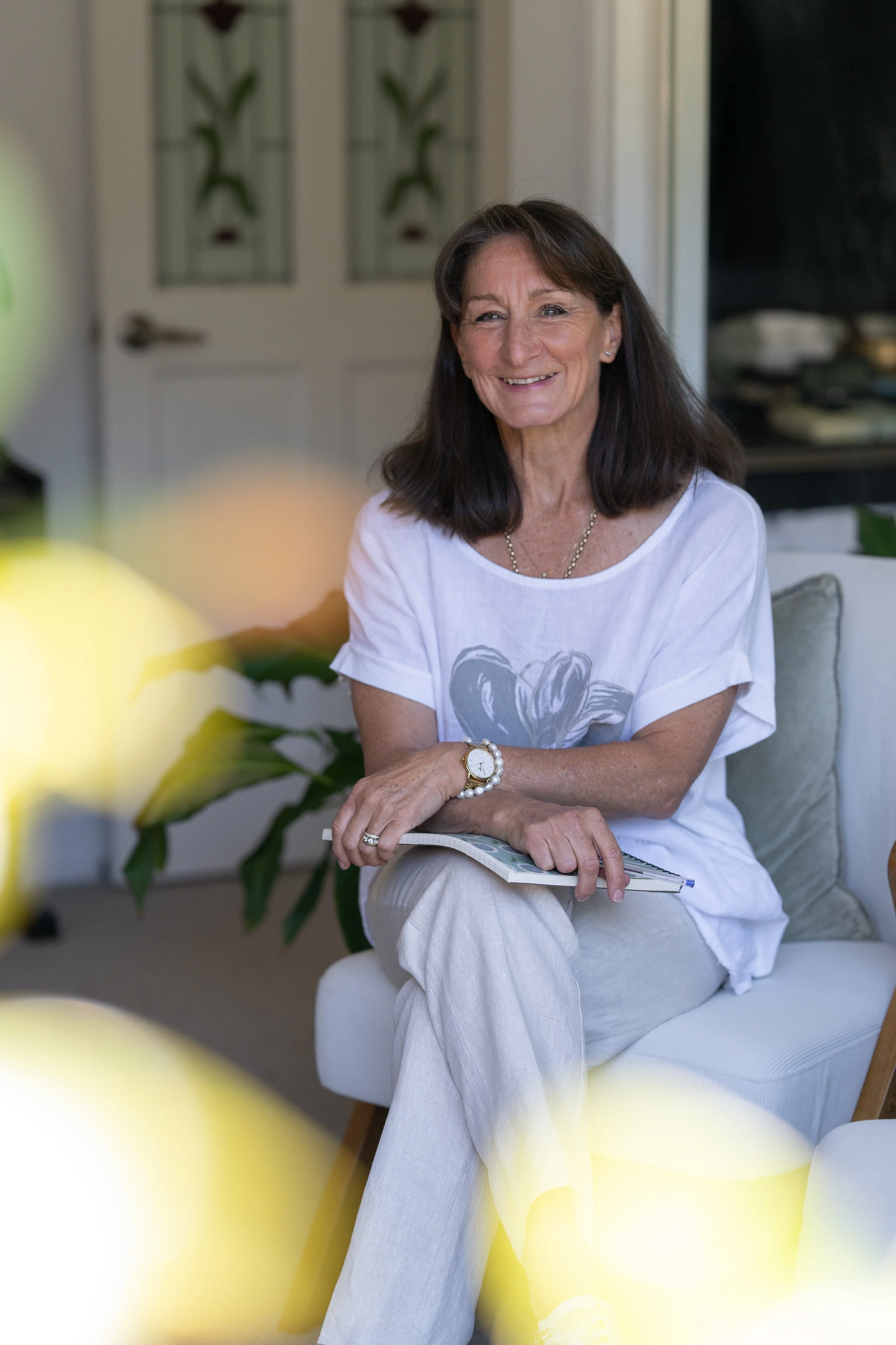 A woman with dark shoulder-length hair smiling while sitting on a white chair in a cozy room, holding a notebook, with plants in the background and blurred yellow flowers in the foreground.
