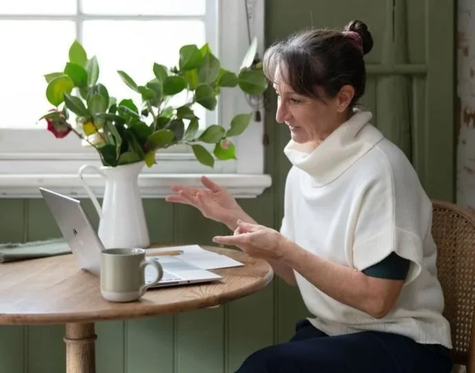 A woman sitting at a wooden table, engaged in a video call on her laptop, with a large vase of roses and a coffee mug on the table. She appears to be smiling or talking outdoors.