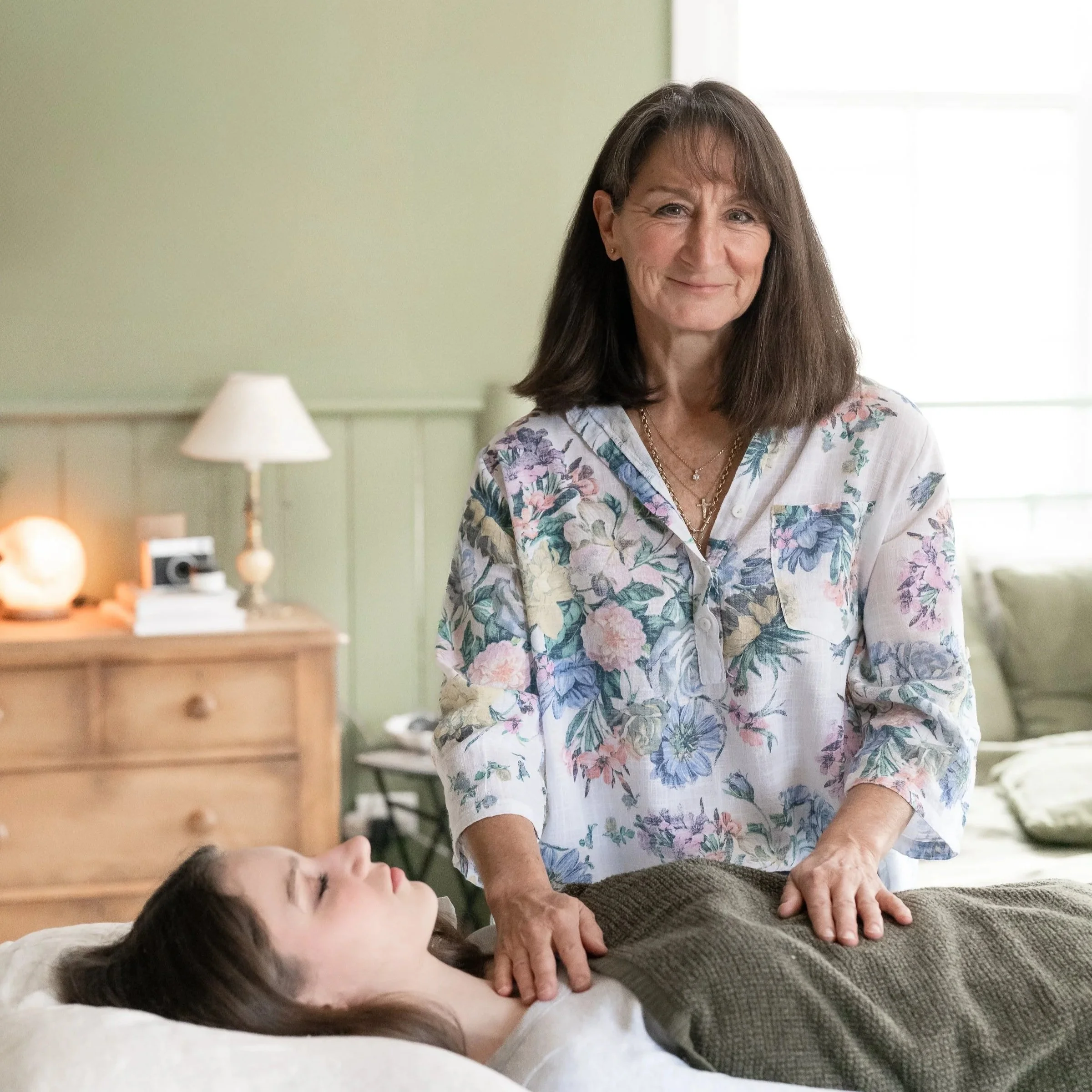 A woman performing Reiki on a girl lying on a treatment bed in a cozy bedroom with green walls, a wooden dresser with a lamp, and a green pillow.