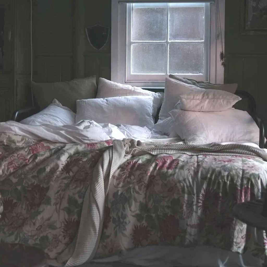 A bed with crumpled white and beige pillows and a floral blanket in a dimly lit room with a window in the background.